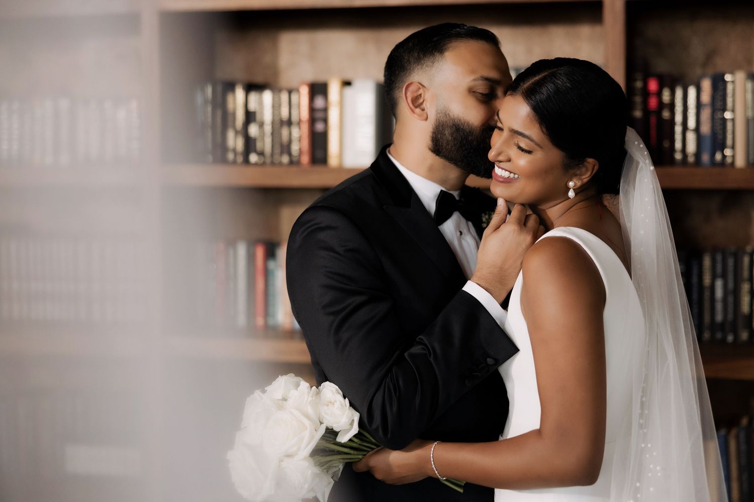Elegant couple in wedding attire embrace near a bookshelf, bride holding white roses in a romantic library setting.