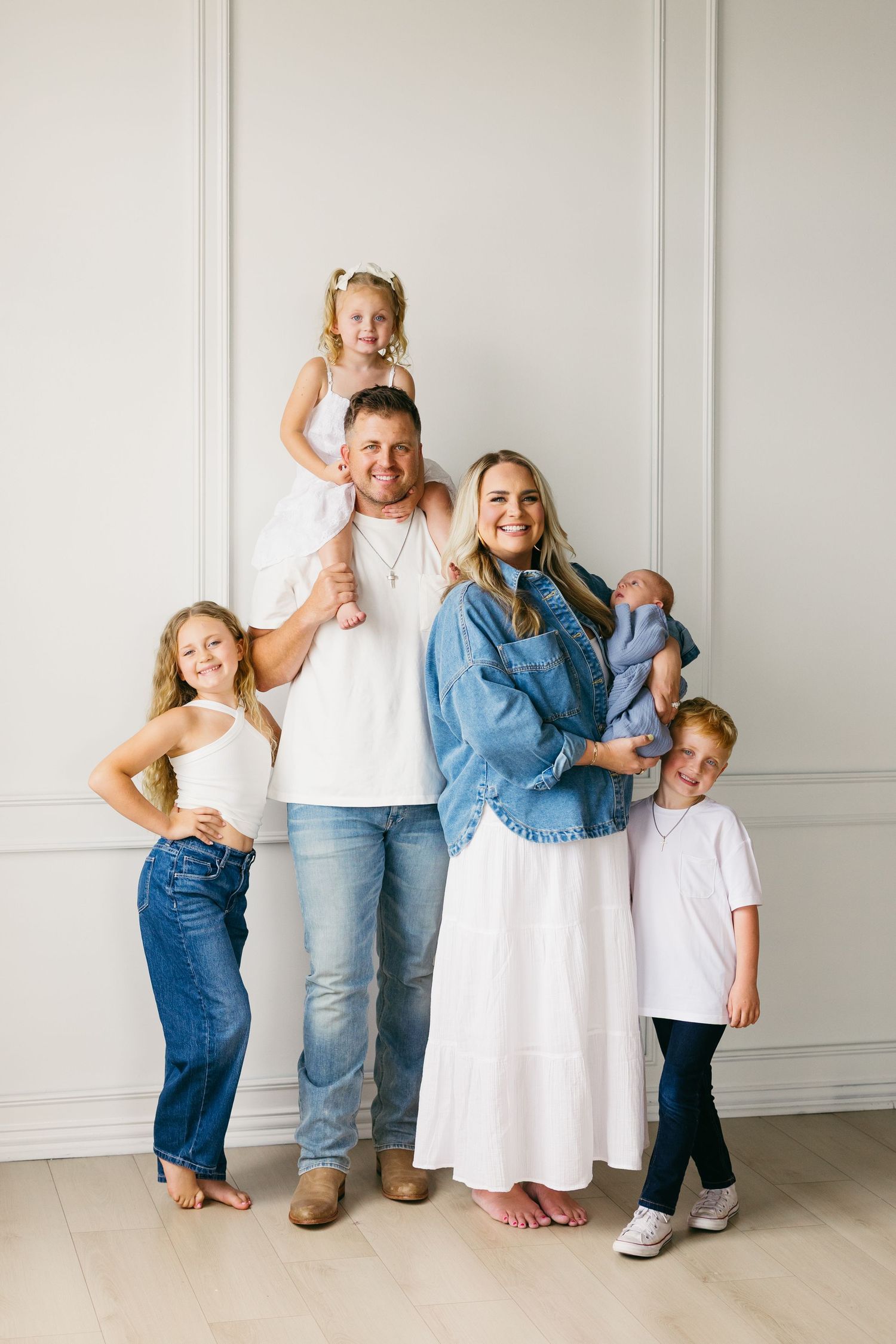 A family poses together against a white wall wearing coordinated casual outfits in white, denim and neutral tones.