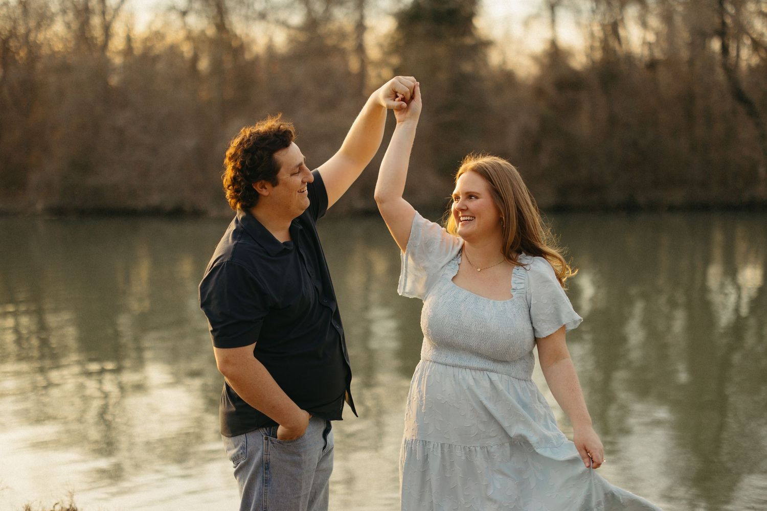 Couple dancing outdoors by a sunlit lake, man spinning woman in a blue dress, golden hour engagement photo.
