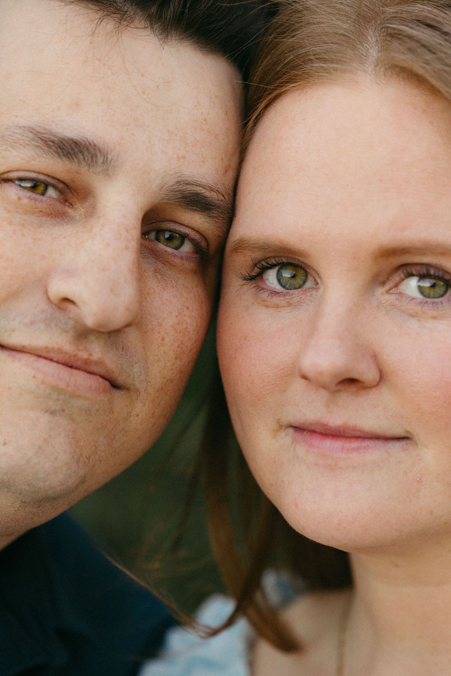 Close-up portrait of a couple with green eyes and fair skin, cheeks touching, smiling softly at the camera.
