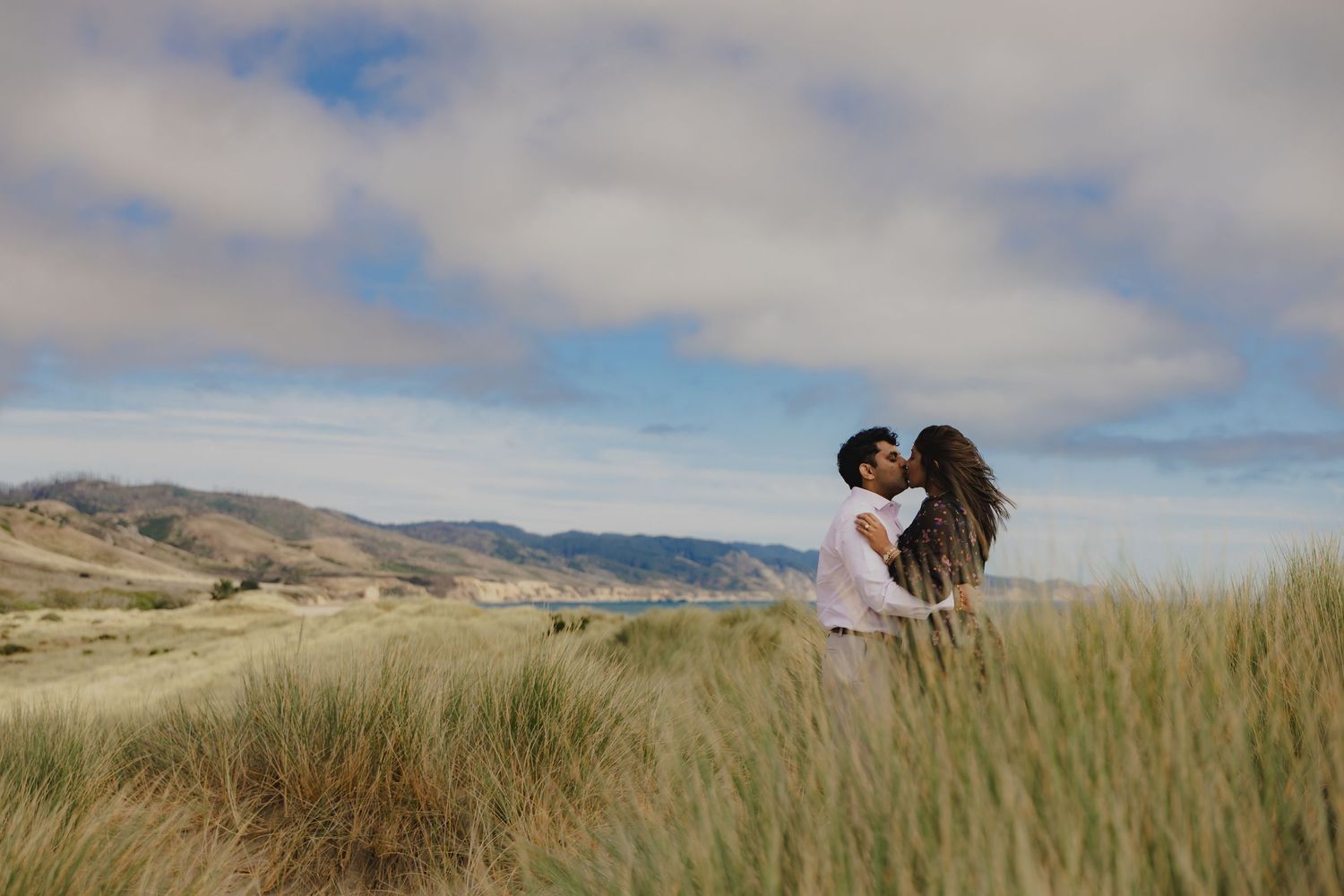 Couple sharing a romantic kiss in tall grass dunes with scenic coastal hills and cloudy blue sky backdrop.
