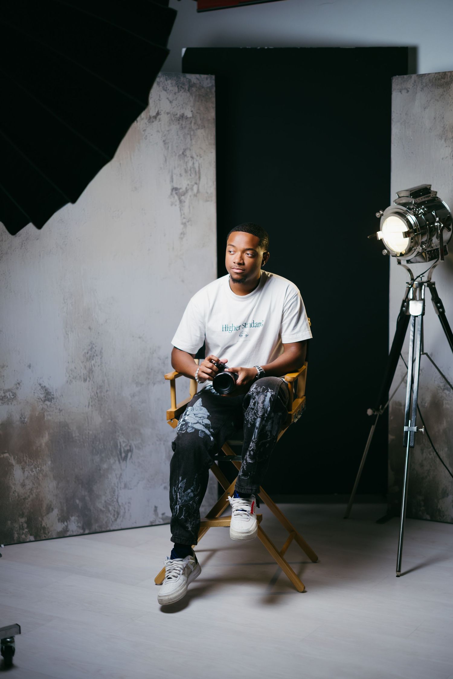 Person sits in director's chair holding camera in photography studio with lighting equipment.
