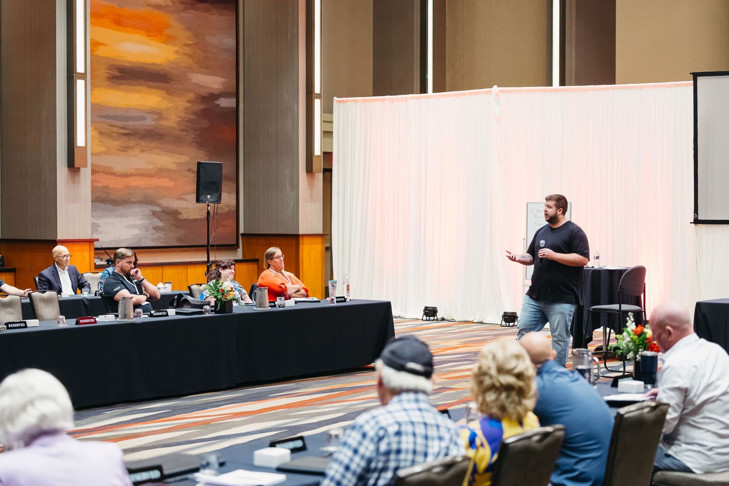 Speaker presents while panel of experts sit at long table during conference session.