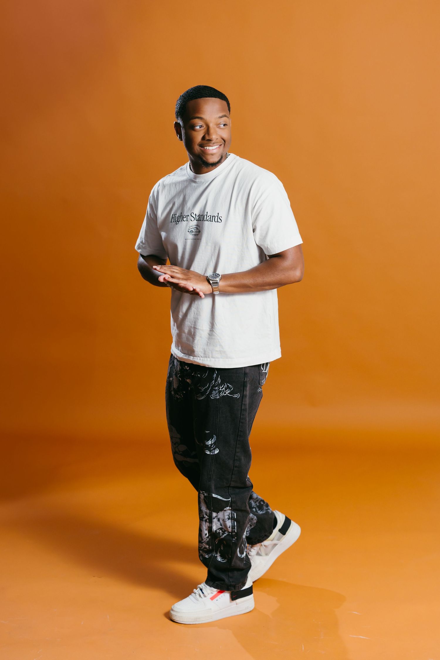 Person in white t-shirt and dark pants poses against orange background with bright smile.