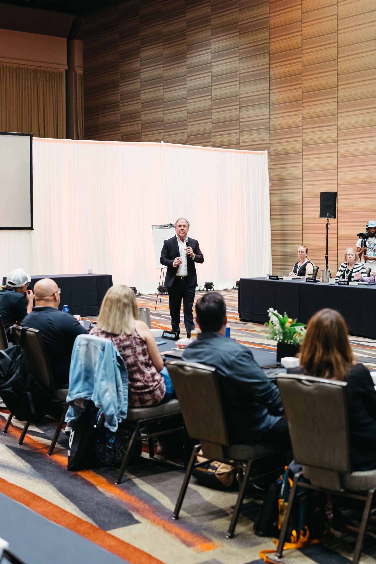 Conference presenter addresses seated audience in modern auditorium setting.