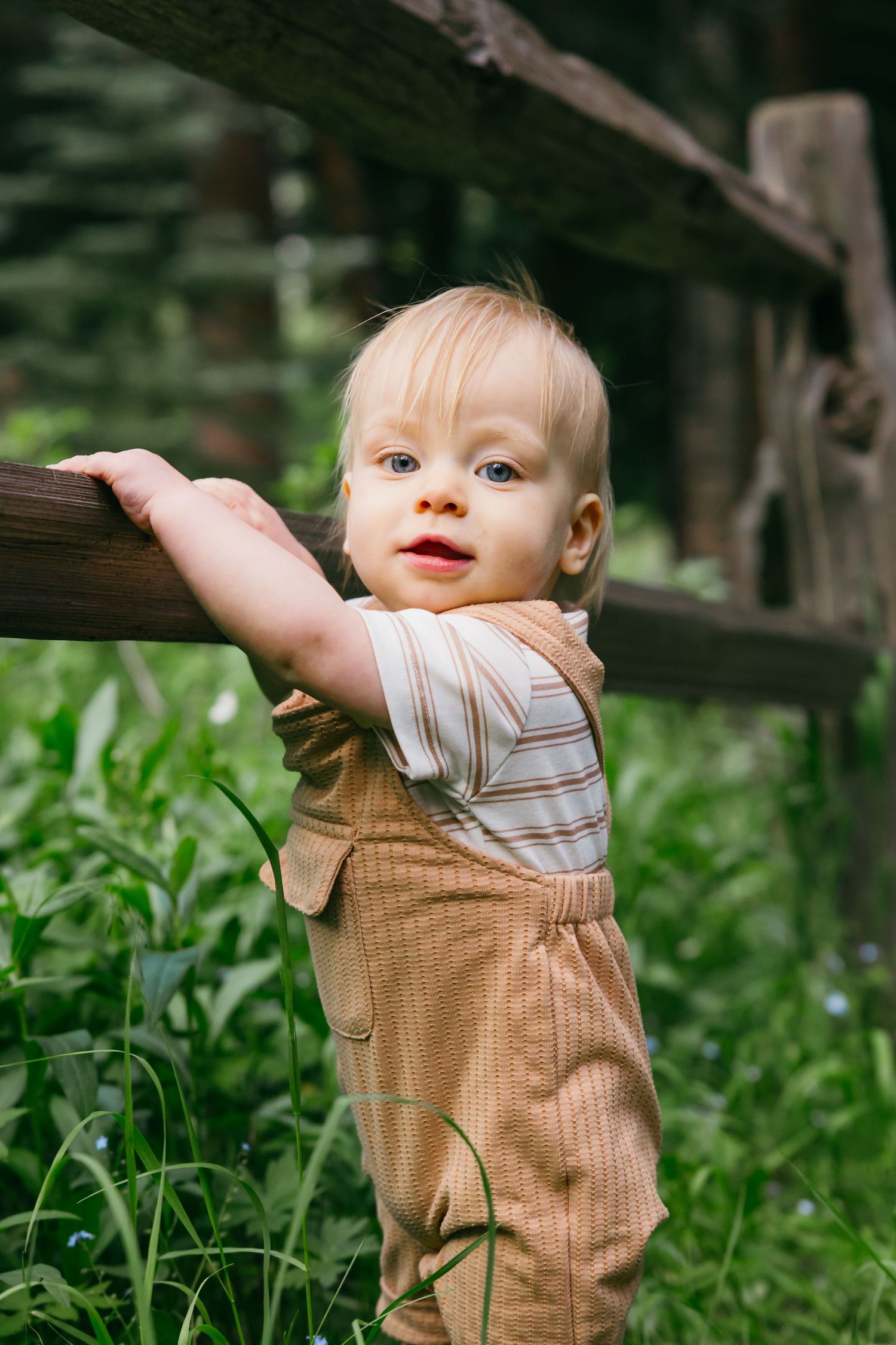 A toddler in brown overalls explores and plays outdoors among green plants.