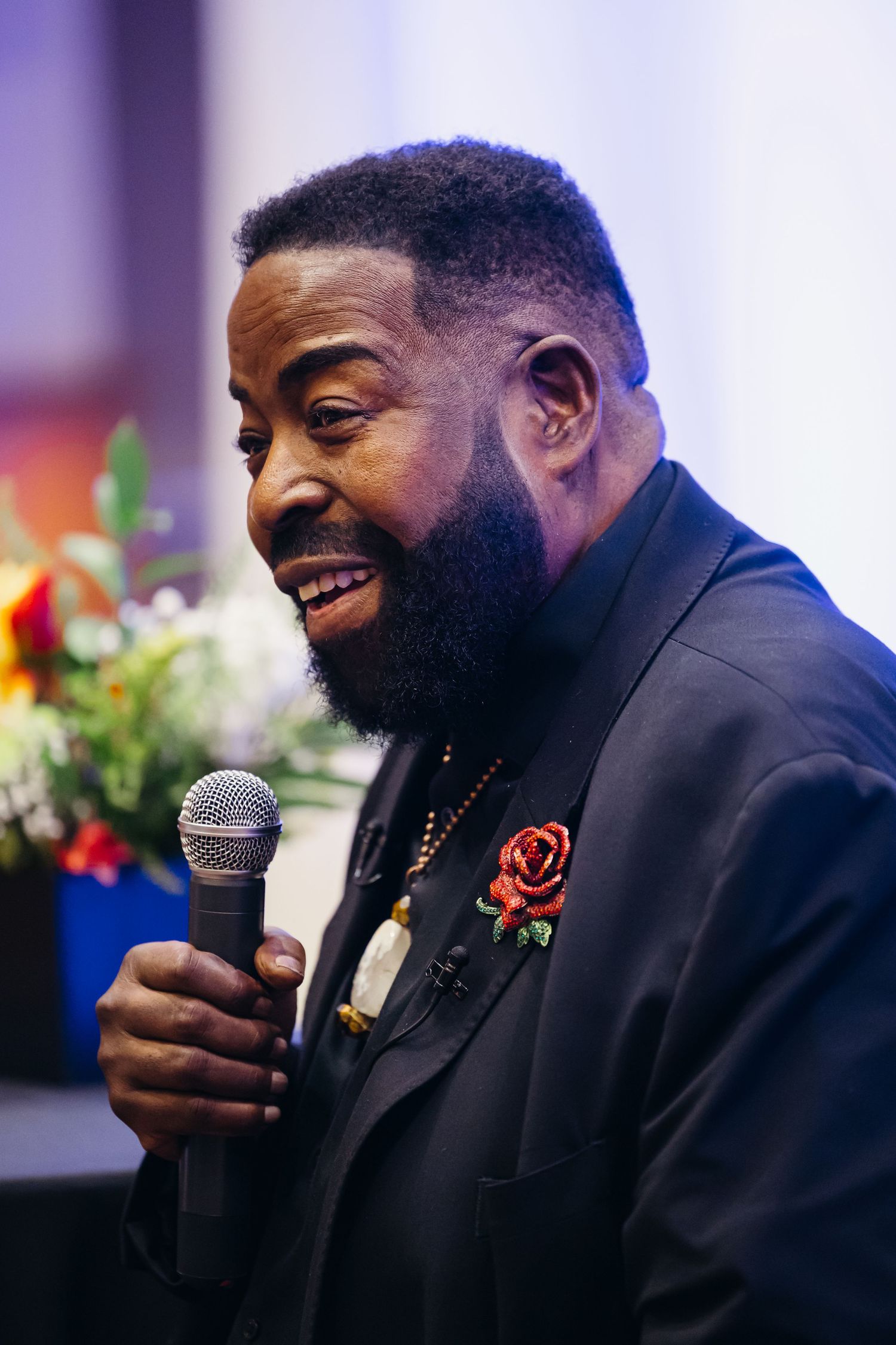 Speaker in a black suit with rose pin addresses audience with microphone at professional conference event.