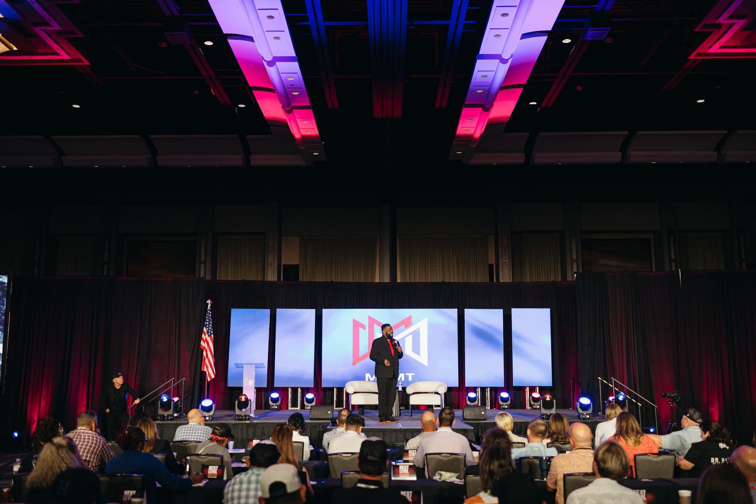 Wide shot of a conference presentation with colorful stage lighting and audience in silhouette.