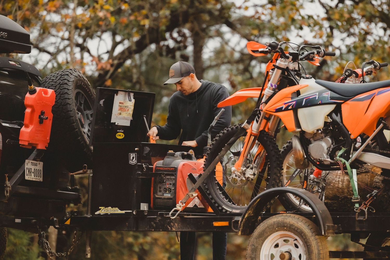 Man loading an orange KTM dirt bike onto a trailer next to a 4x4 vehicle in a wooded outdoor setting.