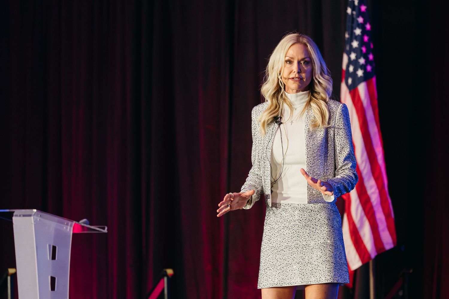 Presenter in light colored outfit speaking at podium with American flag backdrop.