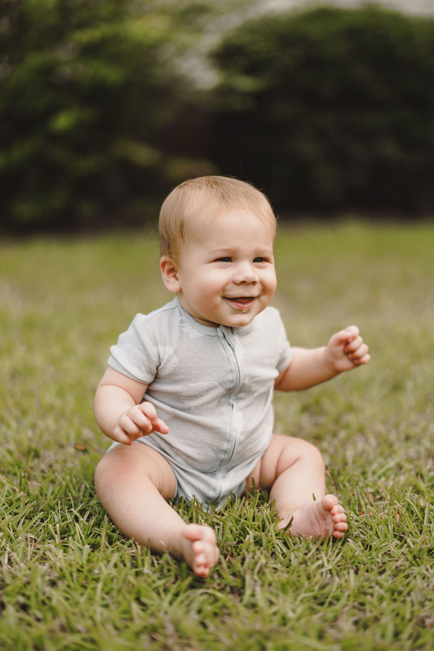 Happy baby in gray onesie sitting on green grass outdoors, smiling and reaching out with one hand.