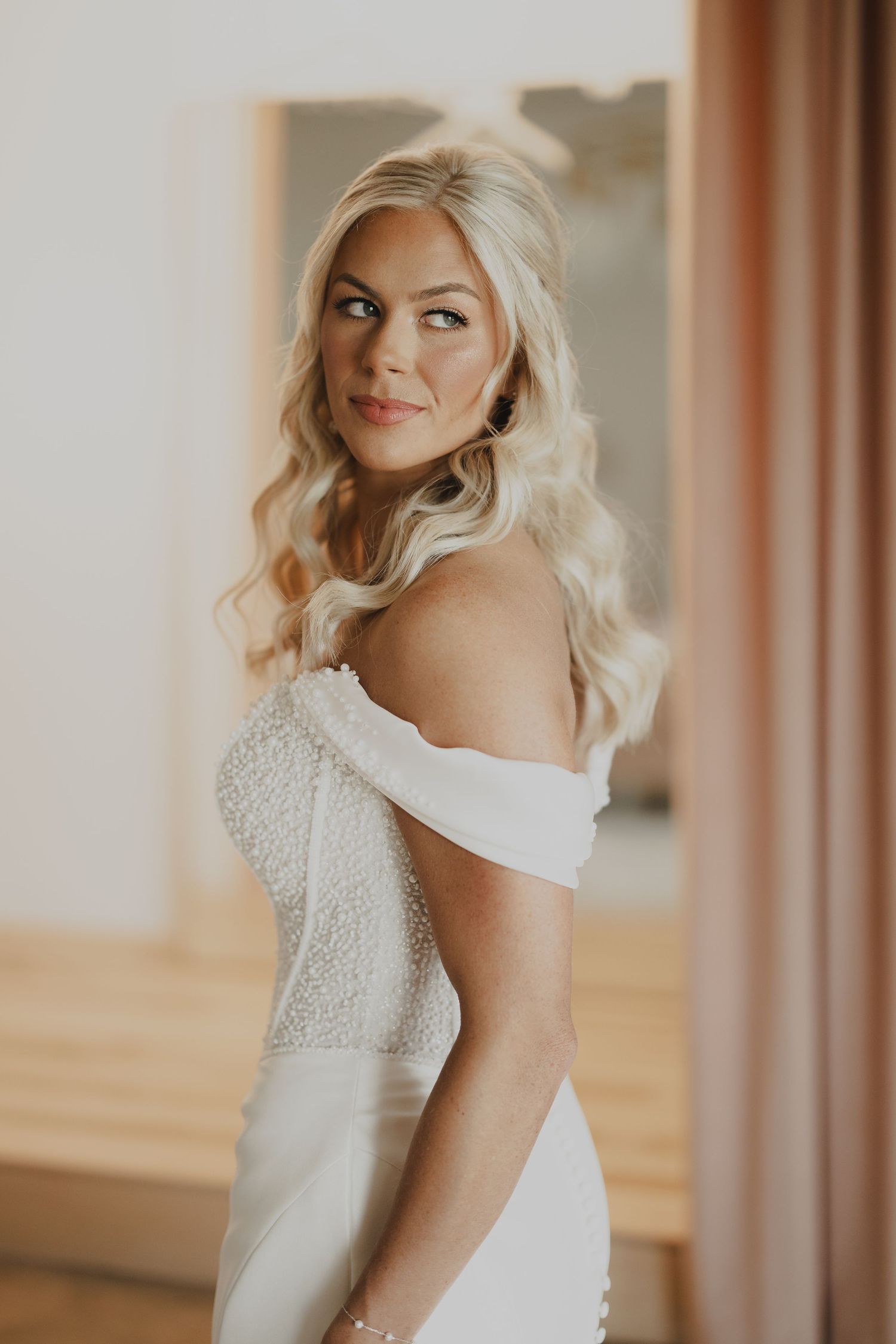 Blonde bride in off-shoulder white lace wedding gown poses elegantly, glancing over shoulder in softly lit room.