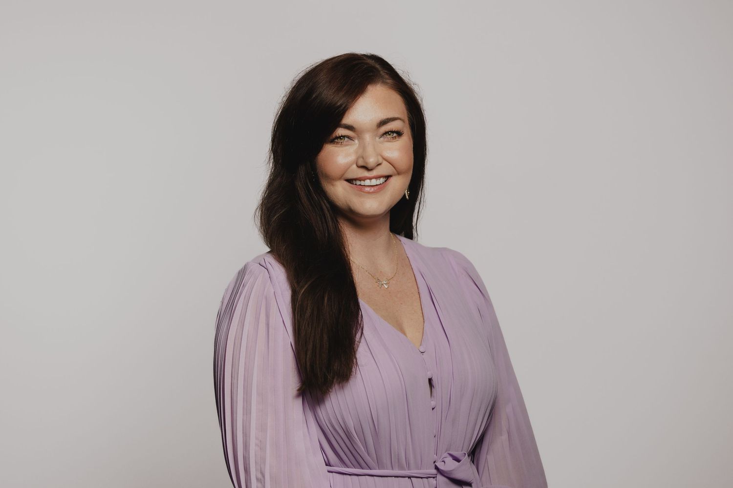 Smiling woman with dark hair wearing a lavender pleated dress, posing against a light gray background.