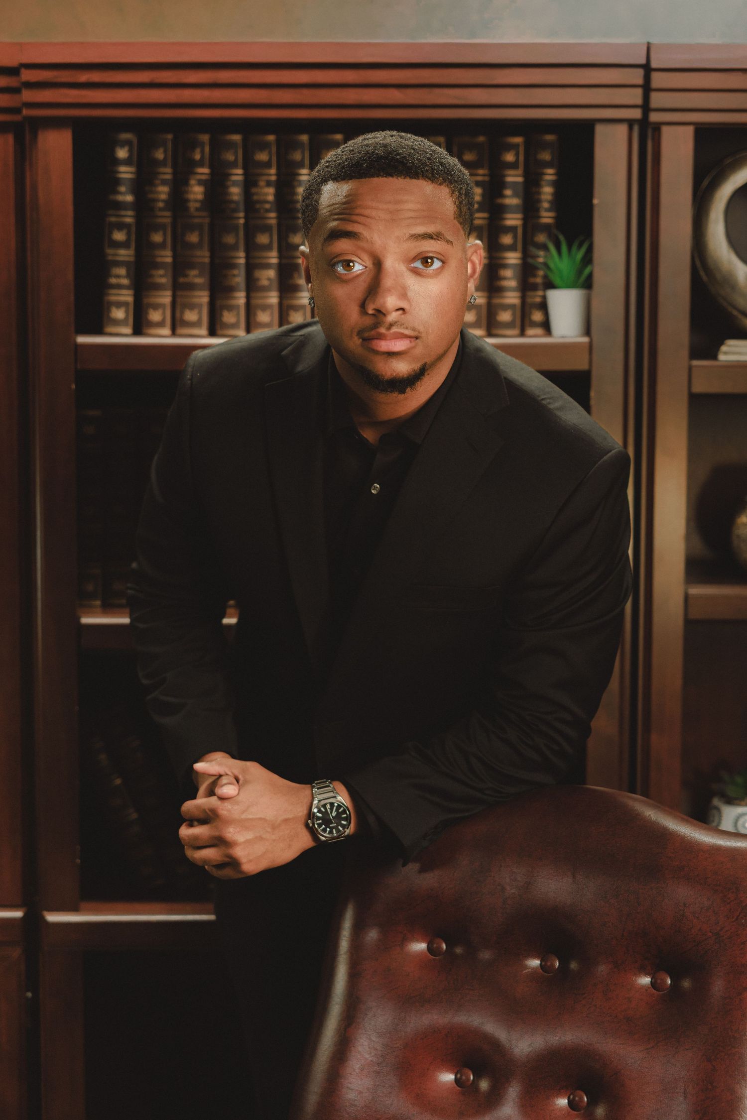 A young Black man in a black suit sits confidently in a leather chair before a bookshelf in a professional setting.