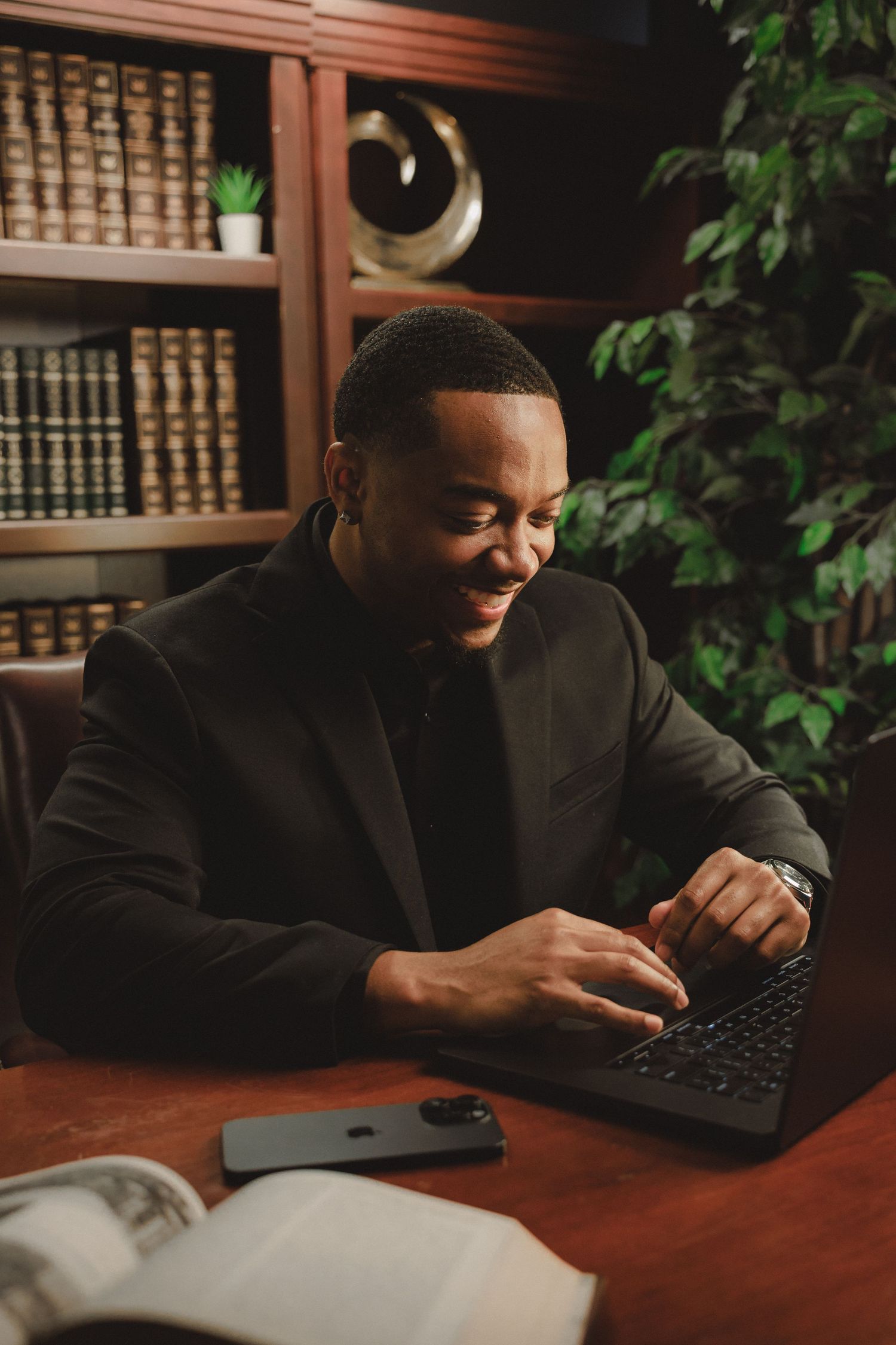 Professional Black man in black suit smiling while typing on laptop in elegant library office setting.