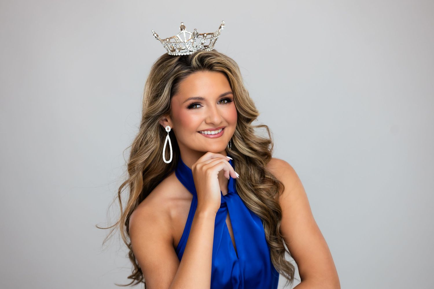Young woman wearing a sparkling tiara crown and blue gown, smiling confidently in a beauty pageant portrait.