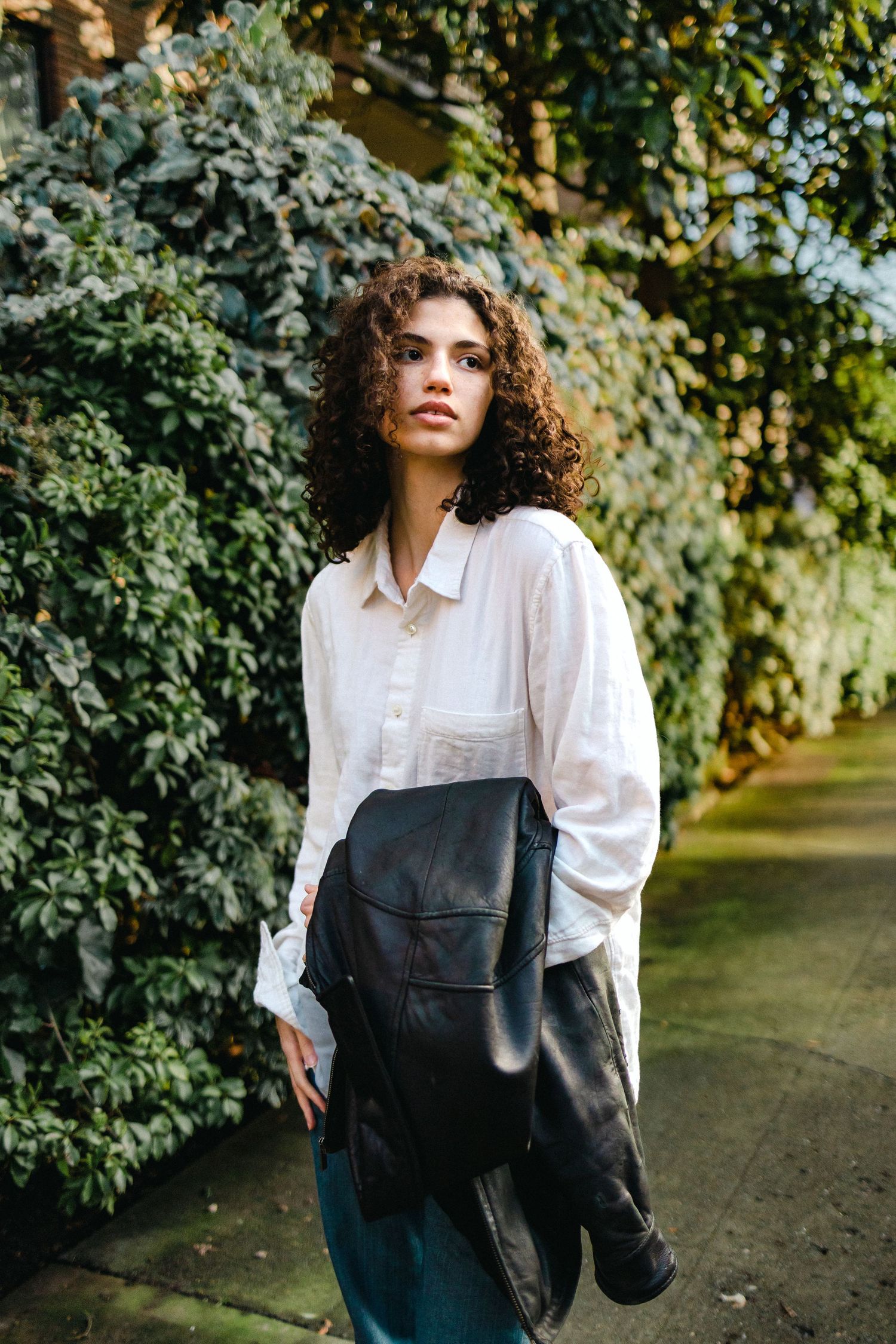 Figure in white shirt and dark overalls poses against green foliage backdrop in outdoor setting.