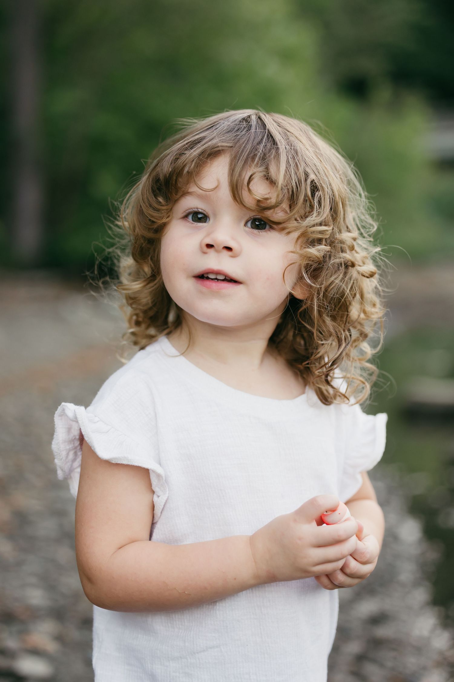 A child in a white dress plays outdoors in a natural setting with trees and foliage in the background.
