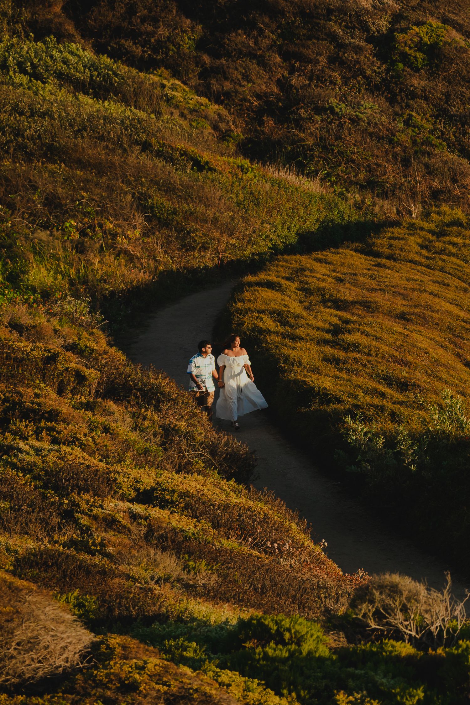 Aerial view of bride and groom walking along a winding coastal trail at golden hour.