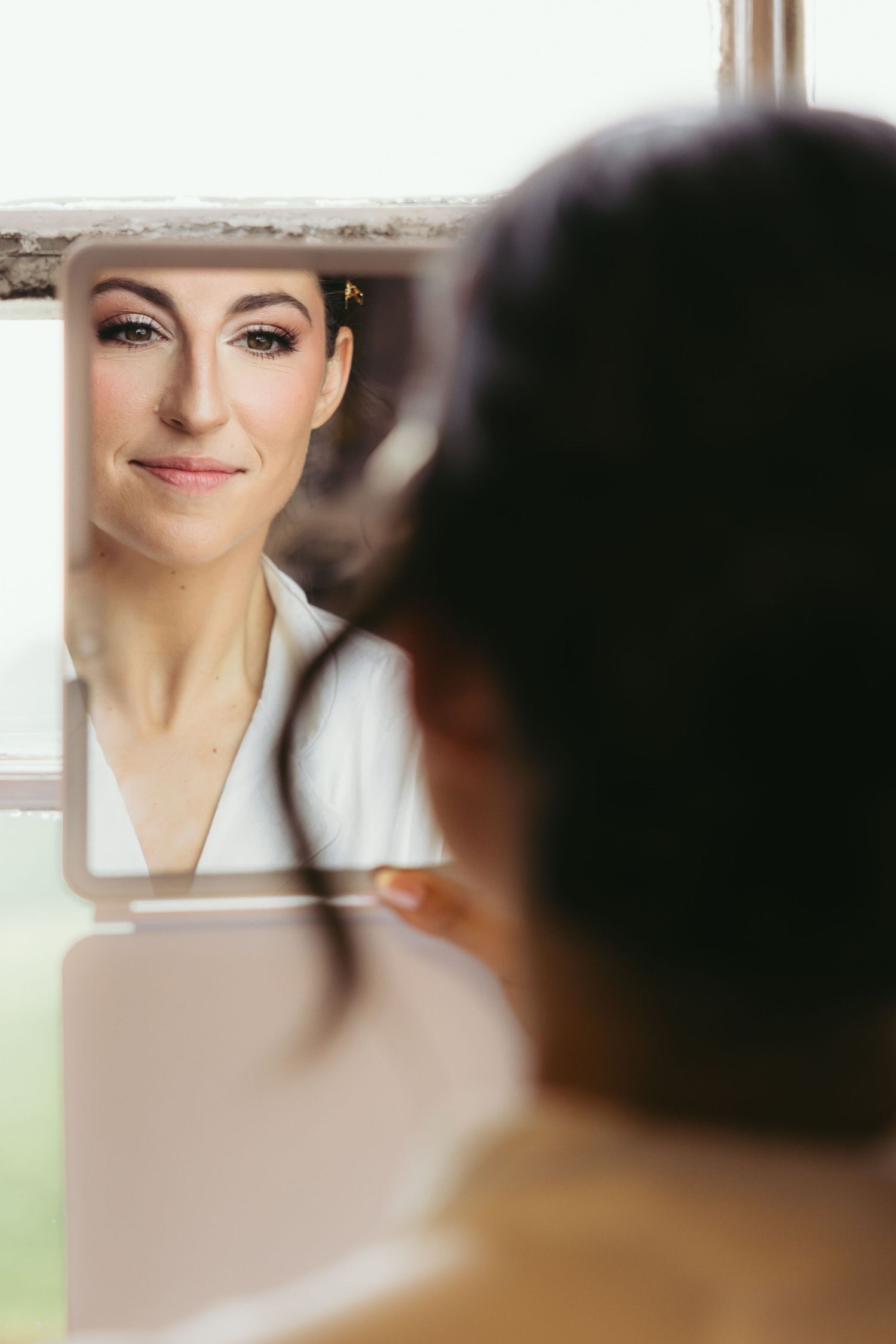 A reflection in a mirror shows a woman wearing a white top with natural makeup and styled hair.