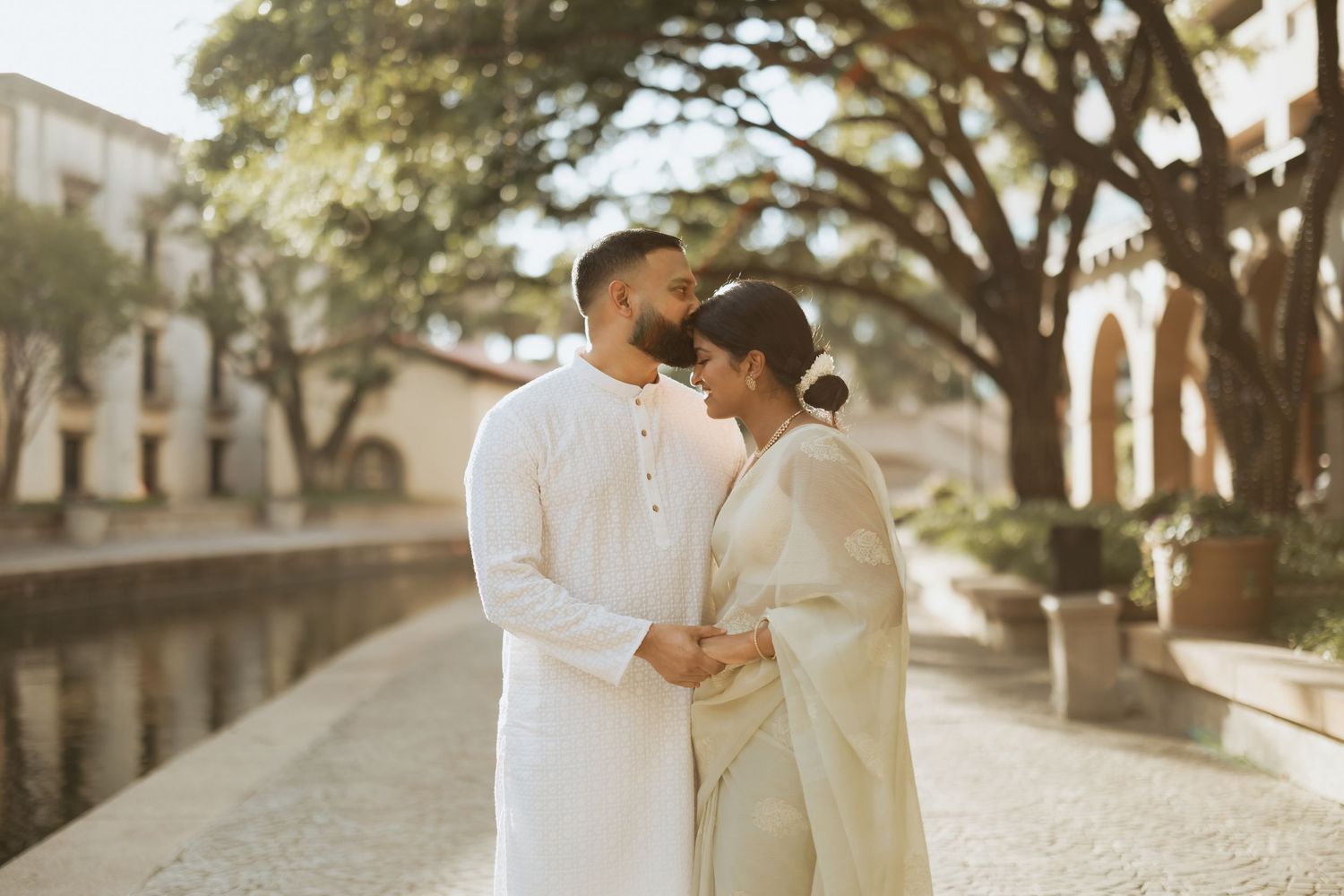 Couple in matching white traditional Indian attire share romantic moment outdoors near waterway.