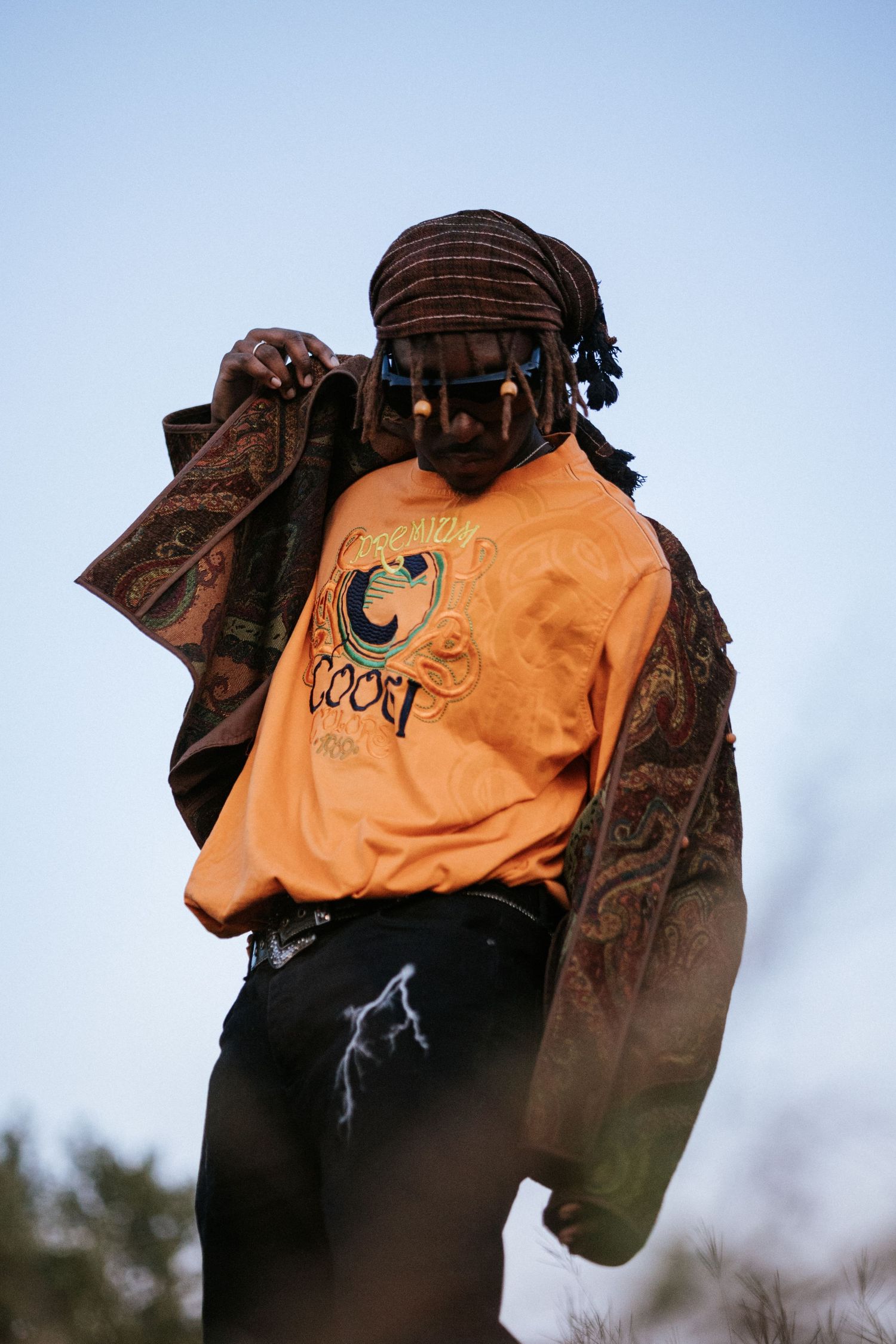 Man in orange graphic tee, plaid jacket, and head wrap posing outdoors against a clear blue sky.