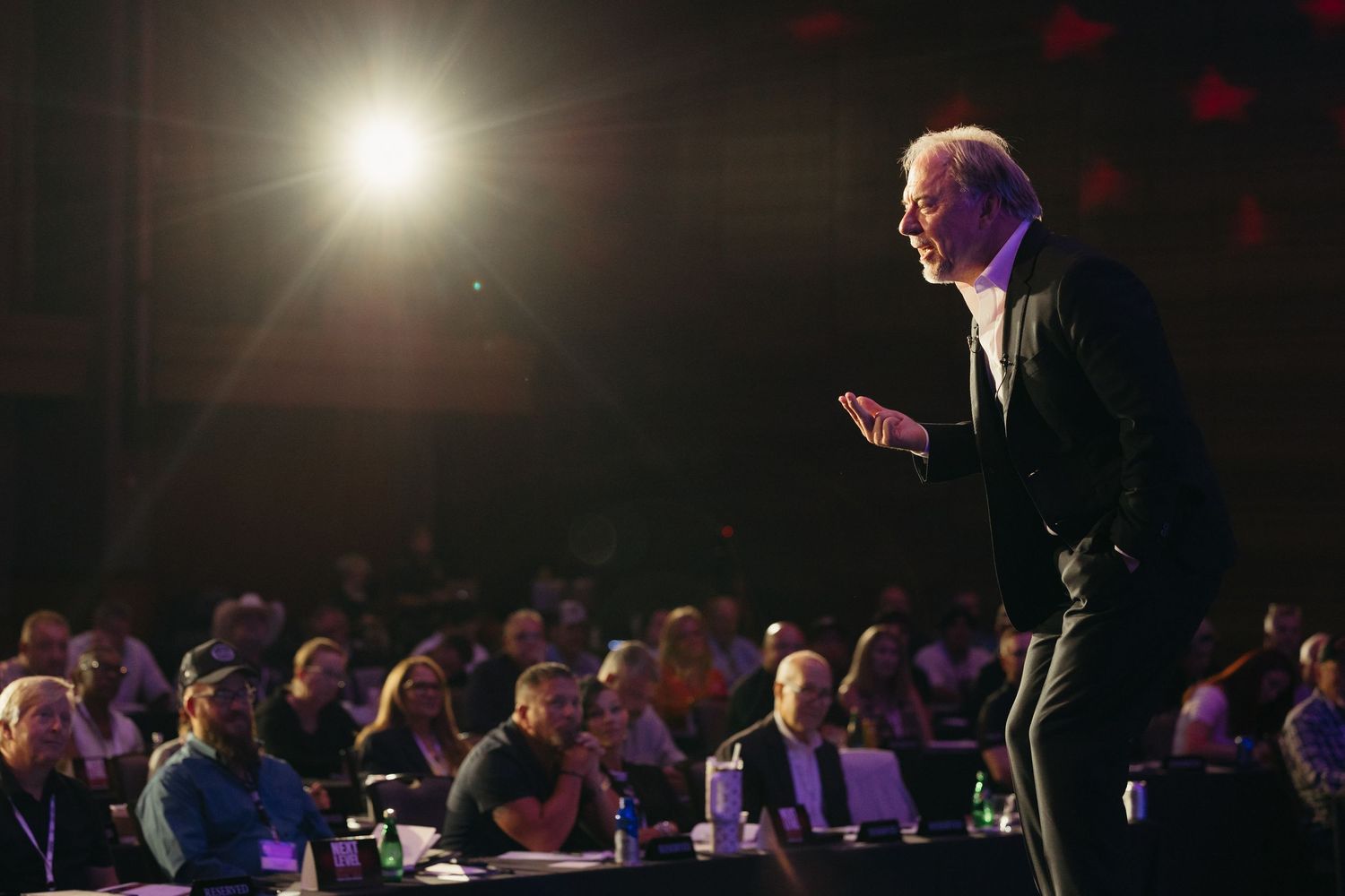 Dramatic stage lighting illuminates speaker presenting to darkened audience.