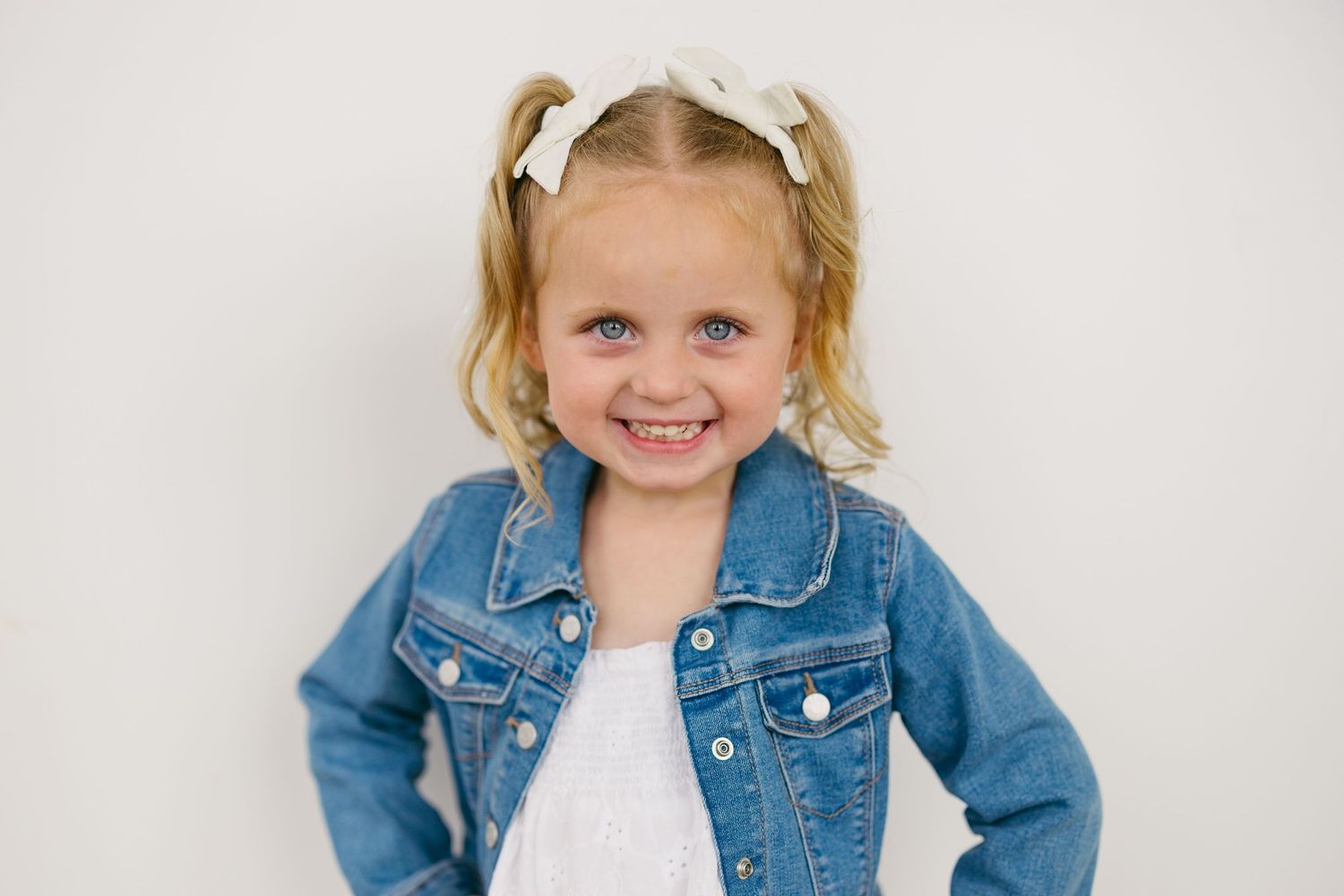 A young child wearing a denim jacket and white shirt poses against a white background with a bright smile.