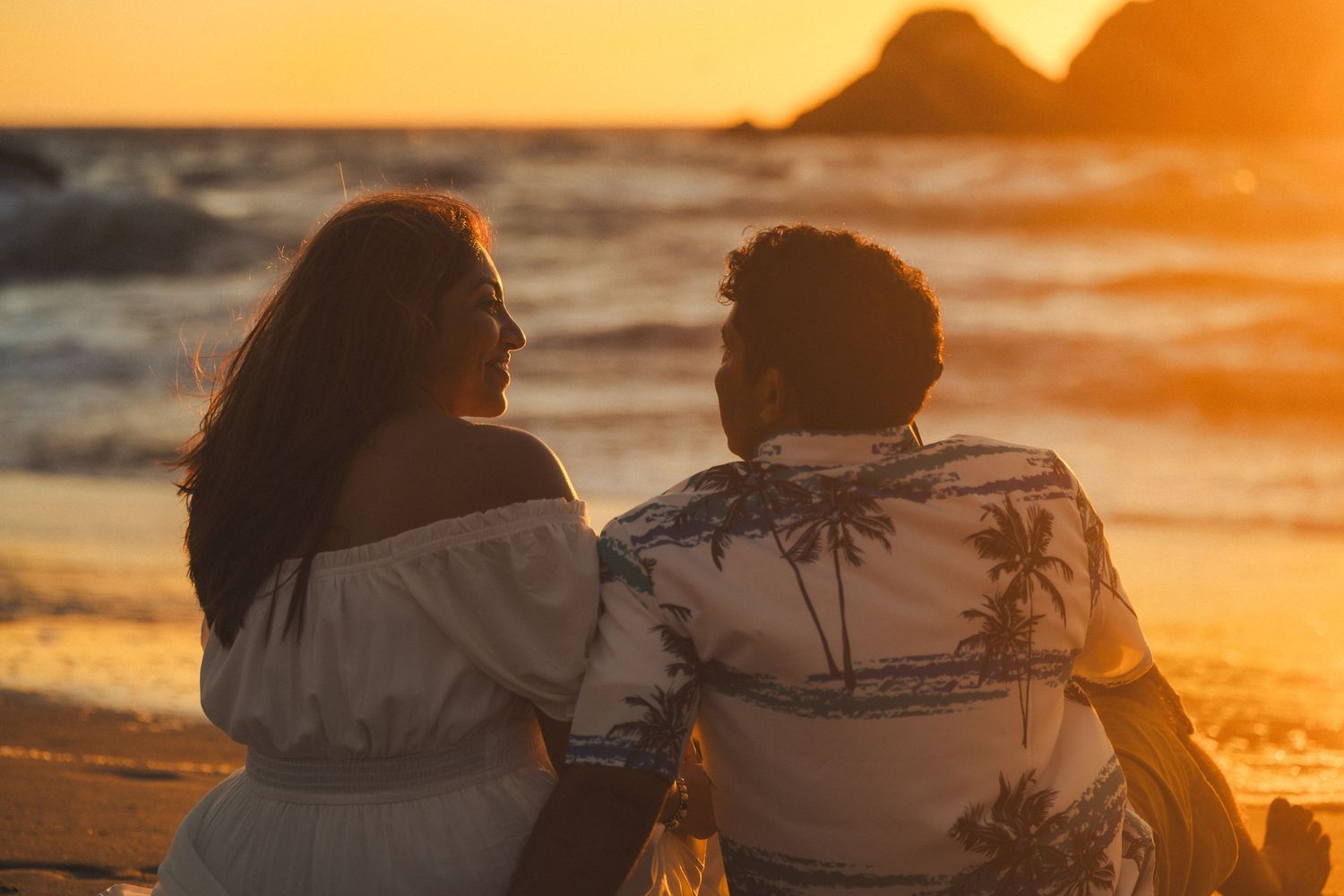 Couple sitting together on a beach at golden sunset, viewed from behind, wearing summer attire with ocean waves.
