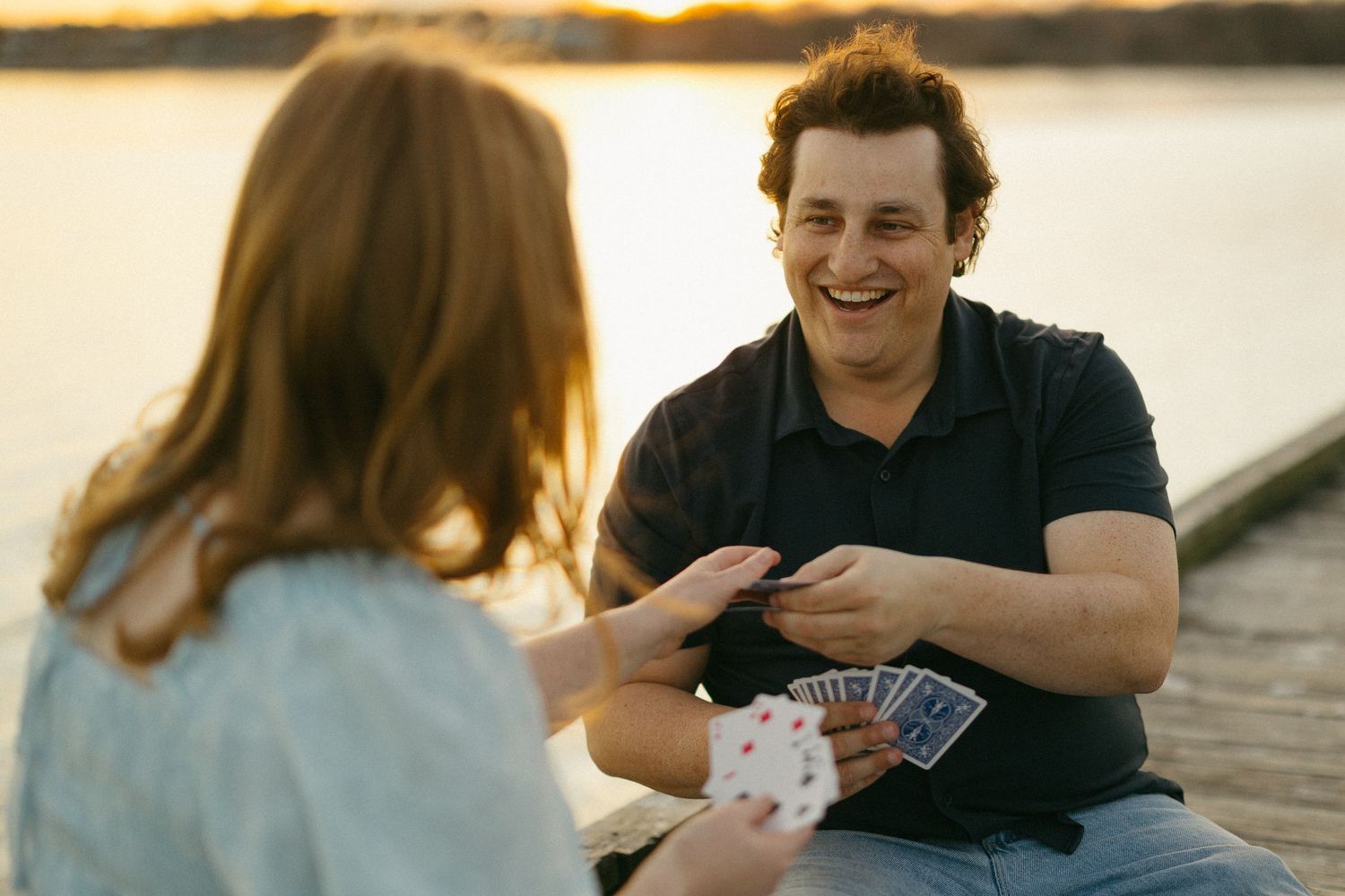 Two people playing cards outdoors by a lake at sunset, man smiling and holding playing cards.