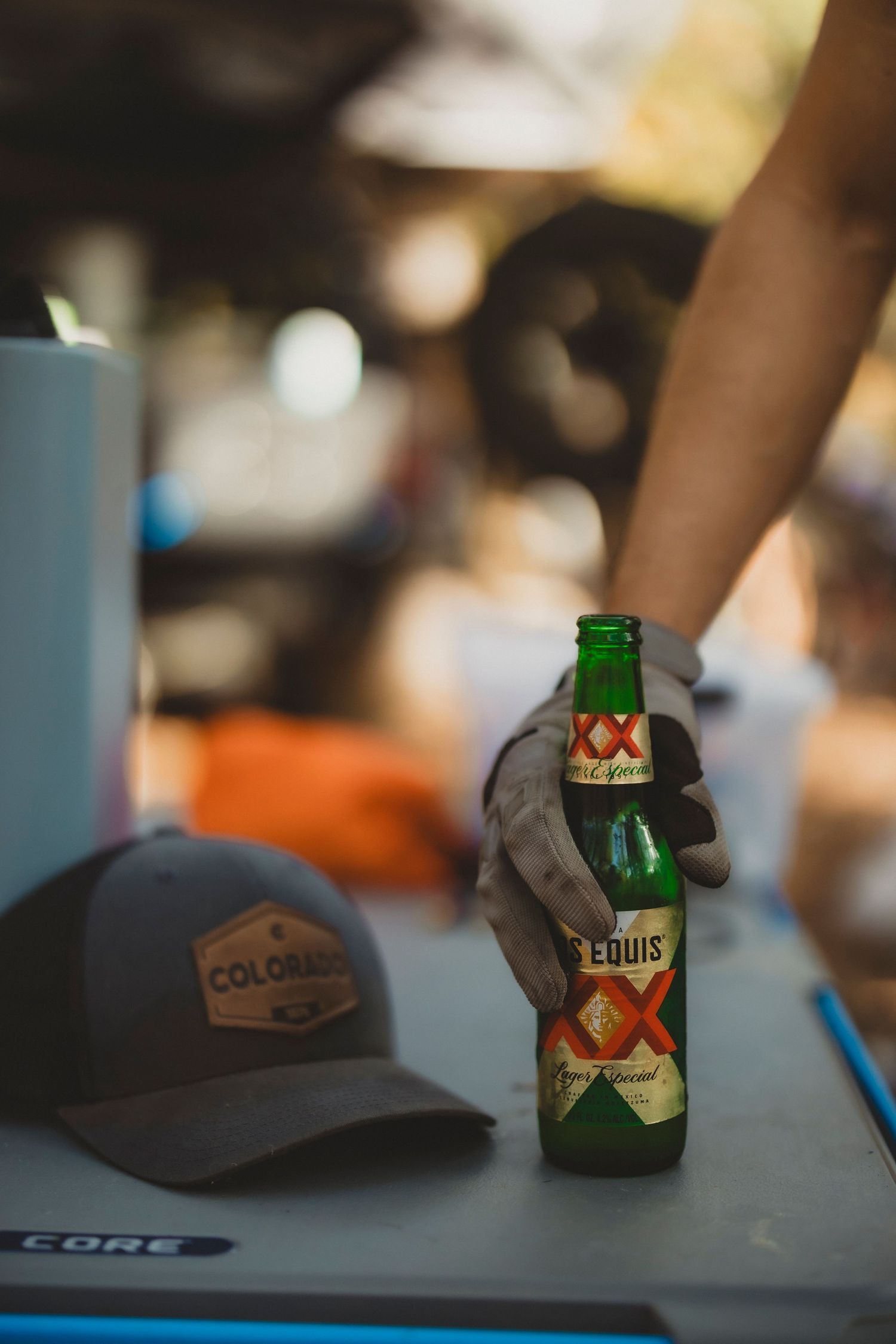 A gloved hand grips a Dos Equis beer bottle outdoors, with a Colorado snapback hat on a cooler nearby.