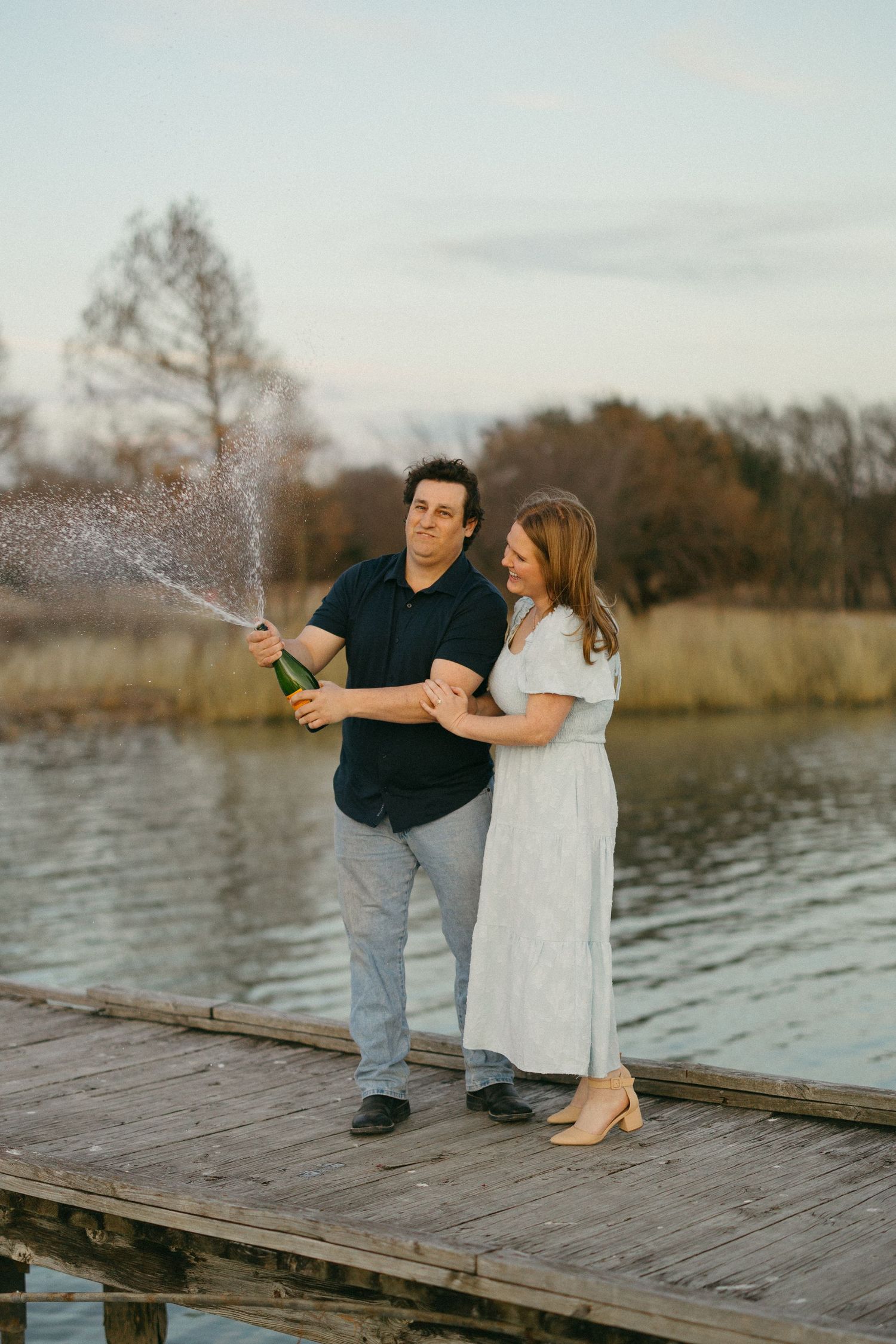Couple celebrating on a wooden dock by a lake, popping champagne bottle together at sunset.