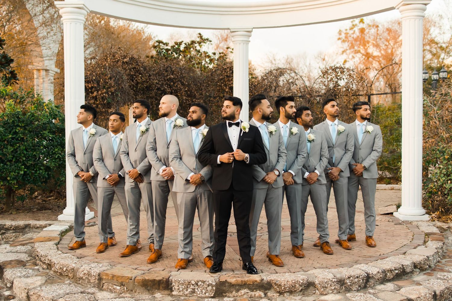 Groom in black suit stands with groomsmen in gray suits and brown shoes under white gazebo outdoors.