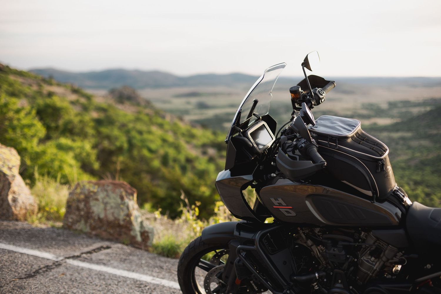 A dark motorcycle parked on a scenic road with lush green hills and a vast valley landscape in the background.