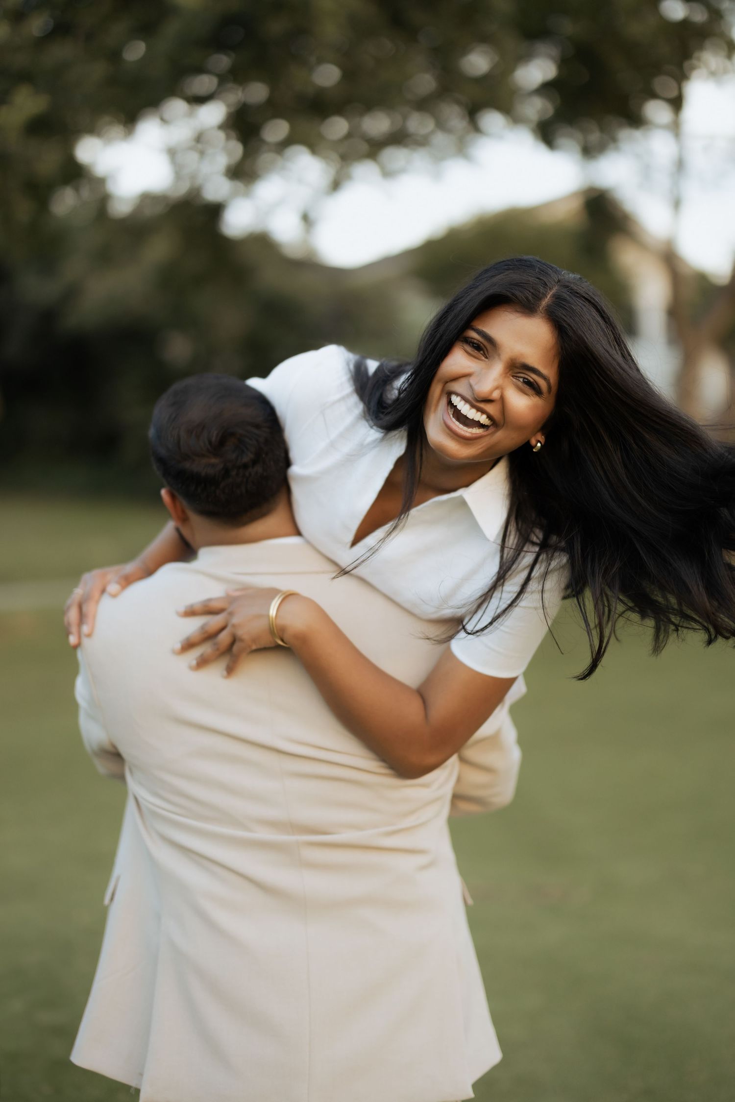 A joyful woman in white laughs while being lifted and hugged by a man outdoors in a lush green park.