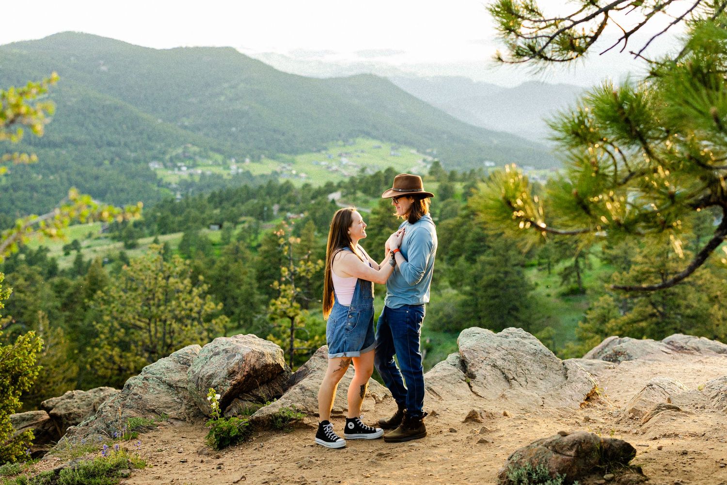 Couple holding hands on rocky mountain overlook with lush green forest valley and hills in background.