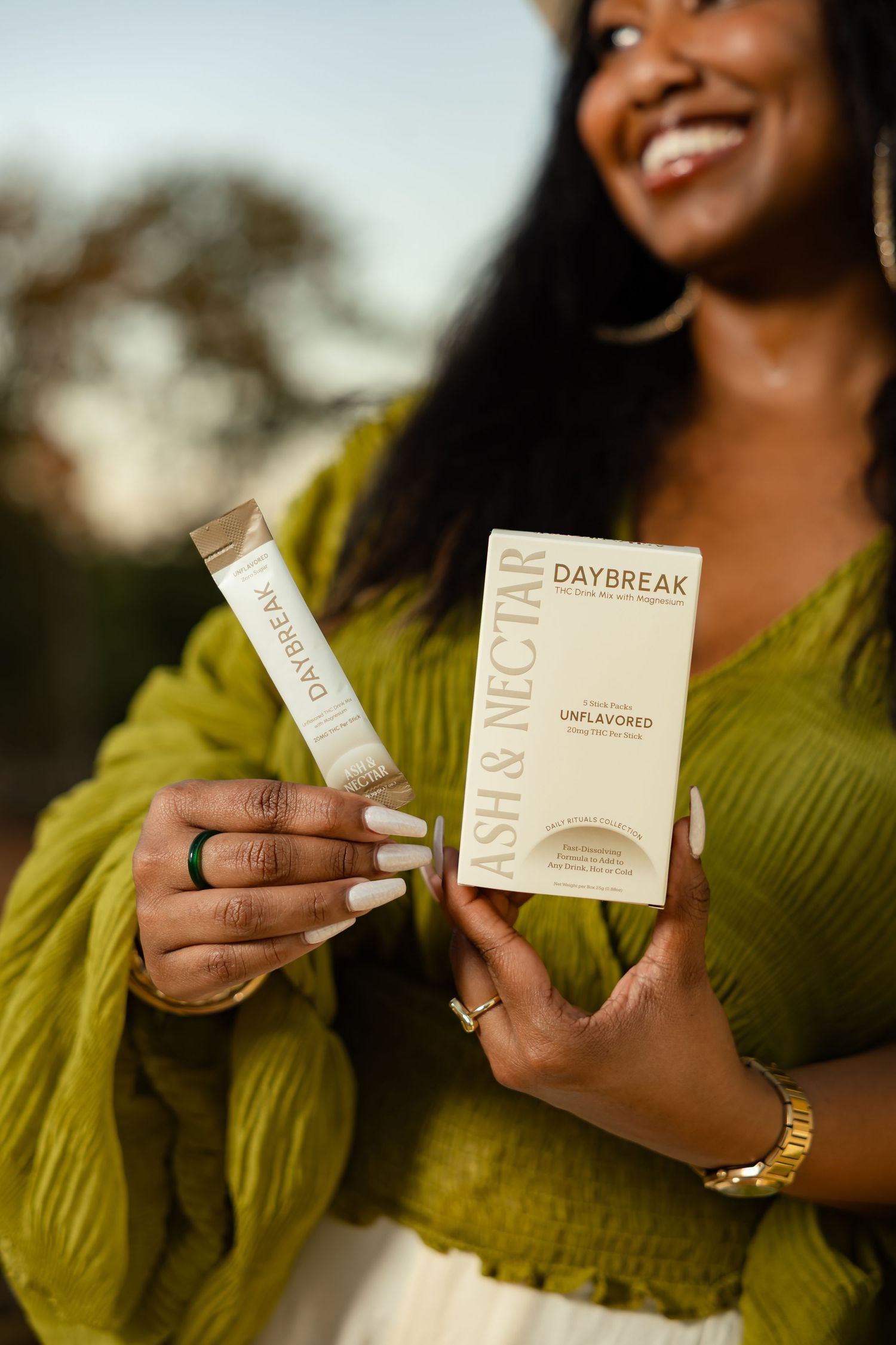 Smiling woman in green sweater holding Ash & Nectar Daybreak unflavored collagen packets outdoors.