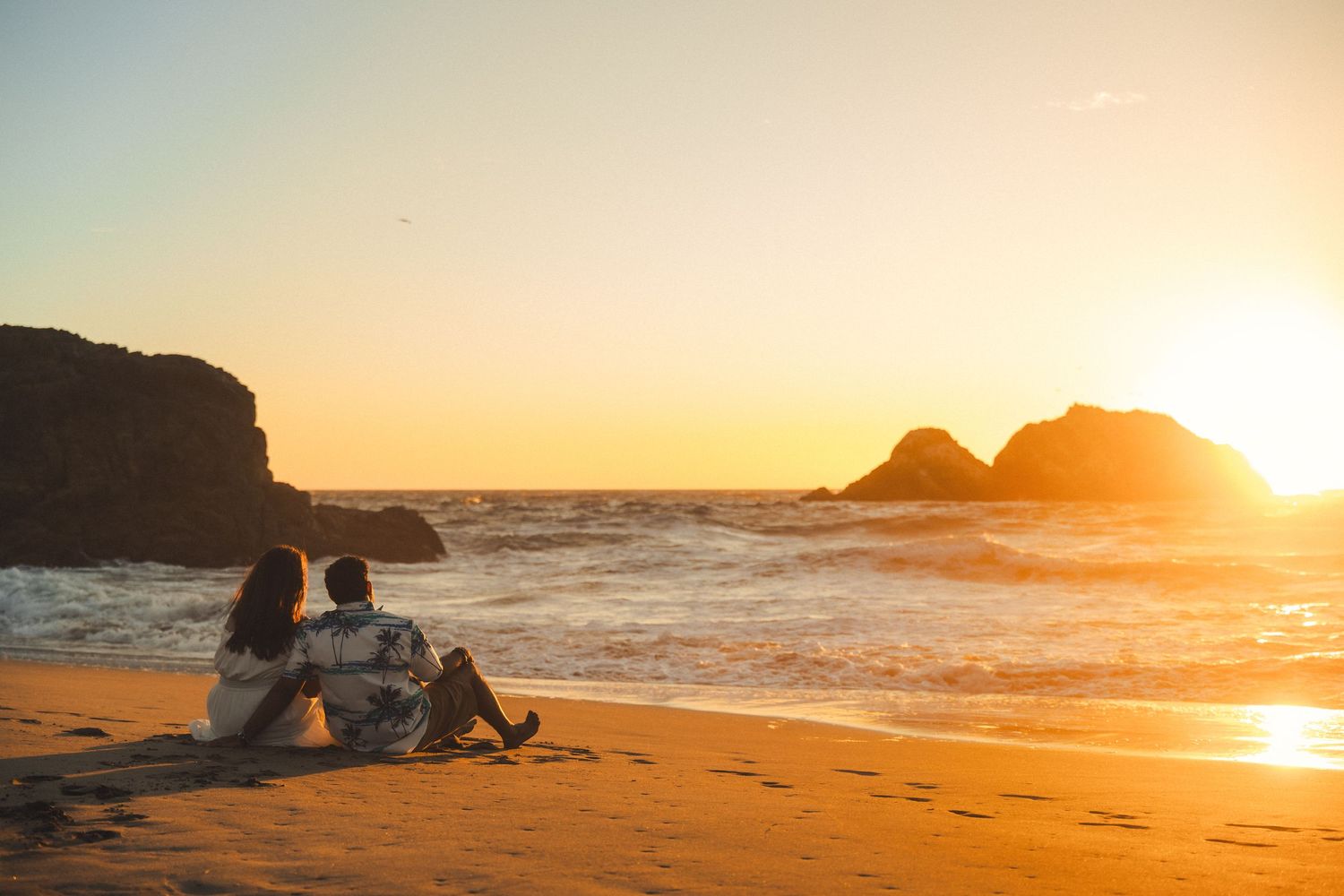 Couple sitting together on sandy beach watching golden sunset over ocean waves with rocky cliffs in background.