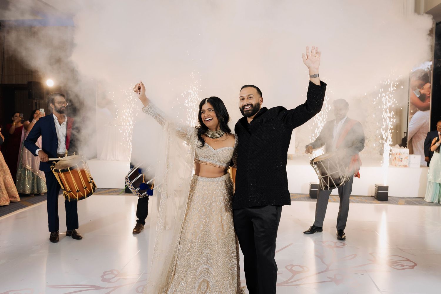 Indian bride and groom celebrate grand entrance at wedding reception with sparklers and dhol drummer.