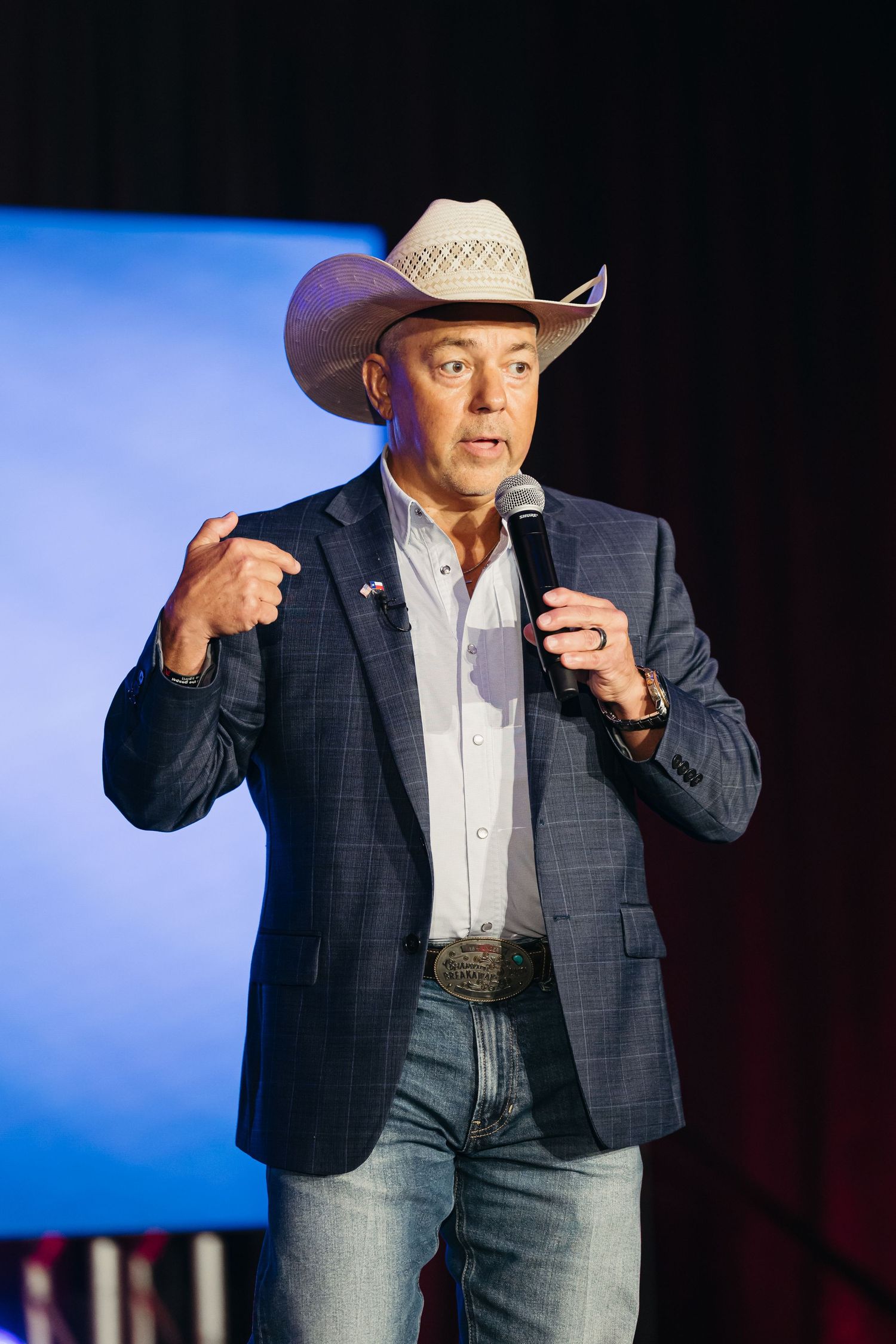 Person wearing a cowboy hat and blazer gesturing while speaking on stage with blue lighting.