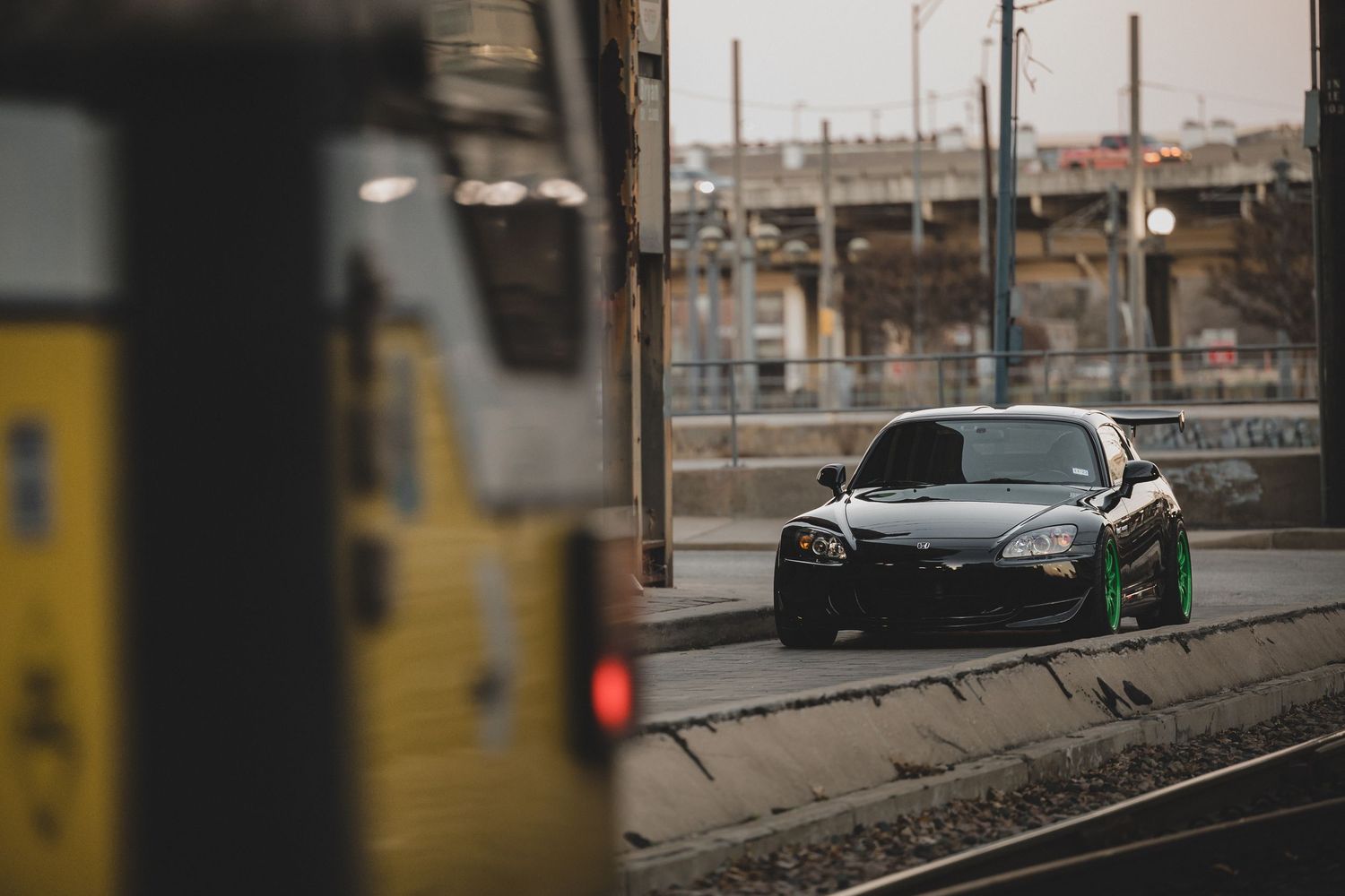 Dark sports car with bright wheels parked near train tracks in an urban setting.