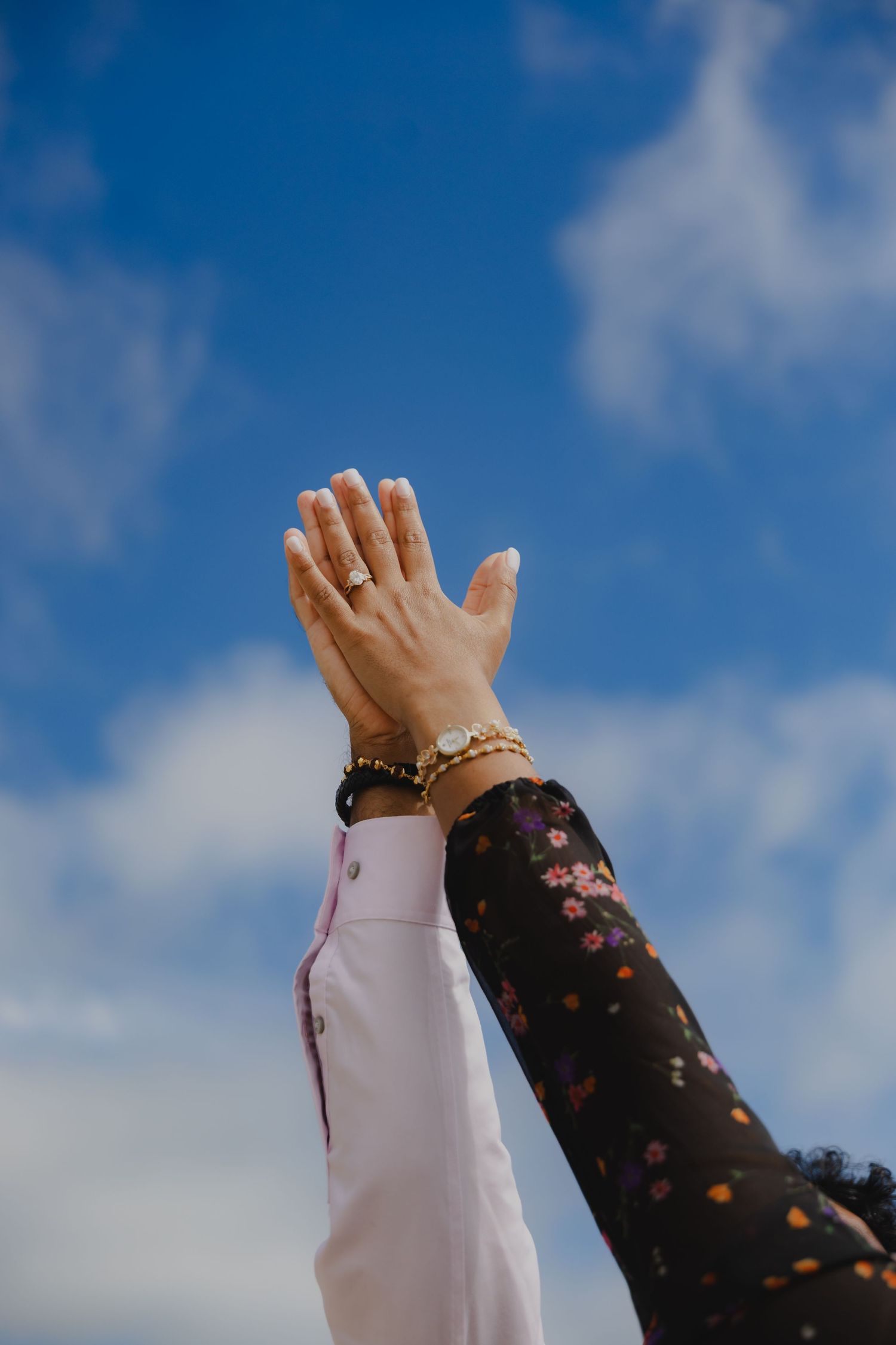Two raised hands high-fiving against a blue sky, one wearing a floral sleeve and gold bracelet, the other in a pink sleeve.