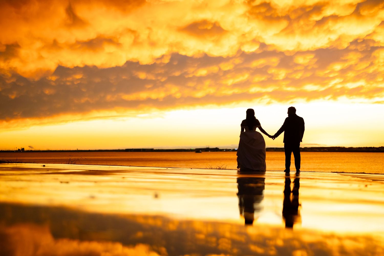 Silhouette of a couple holding hands on a reflective beach at golden sunset with dramatic orange cloudy sky.