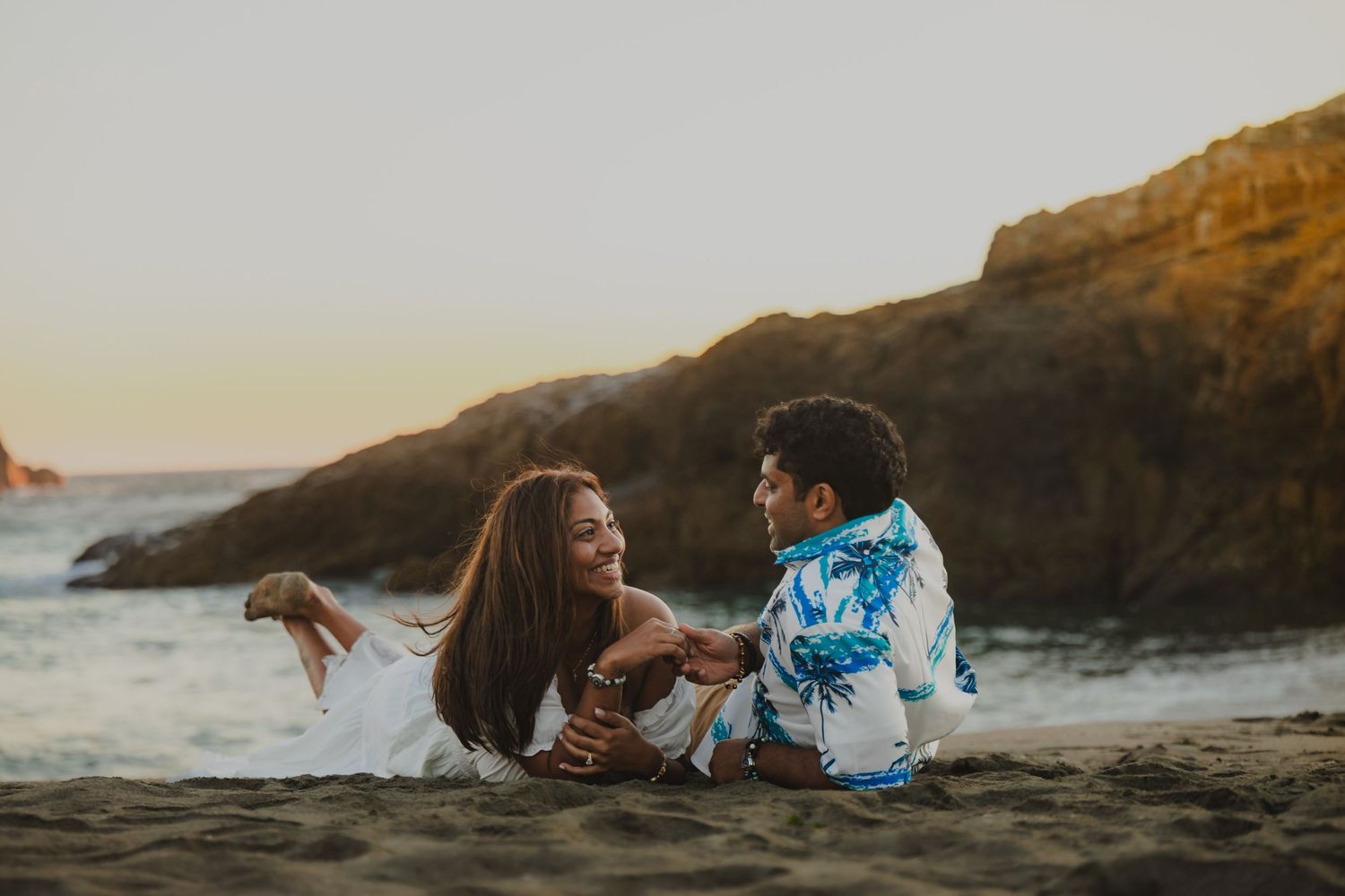 A couple laughs together while lying on a sandy beach at sunset with rocky cliffs in the background.