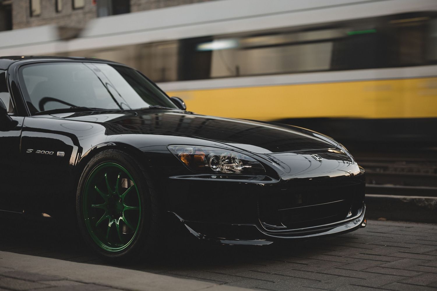 Black Honda S2000 sports car with green wheels parked on a city street with a yellow tram passing in the background.