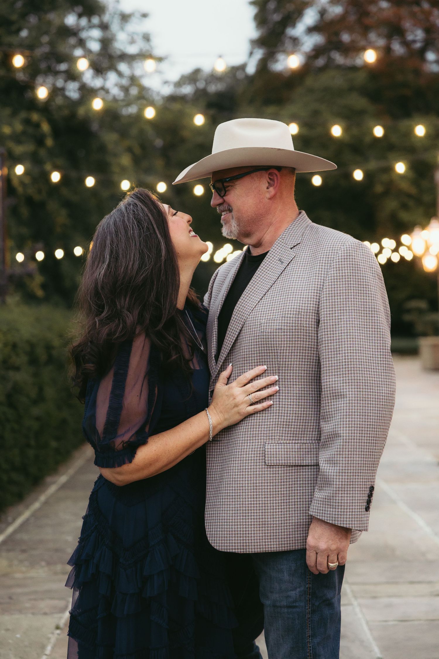 A couple embraces romantically under string lights at dusk, with the person wearing a cowboy hat and checkered blazer.