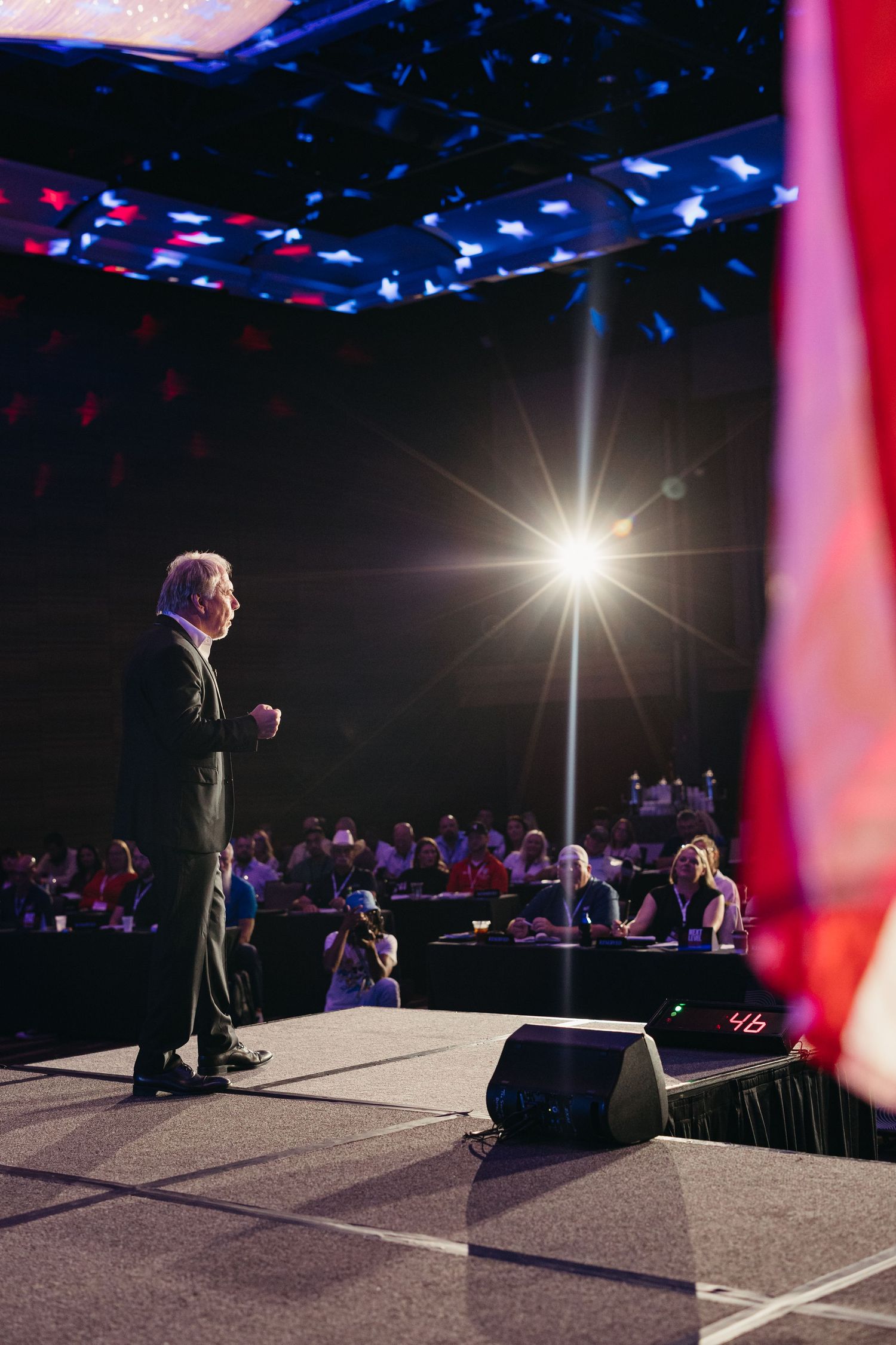 Dramatic stage lighting illuminates presenter speaking to audience in dark conference hall.