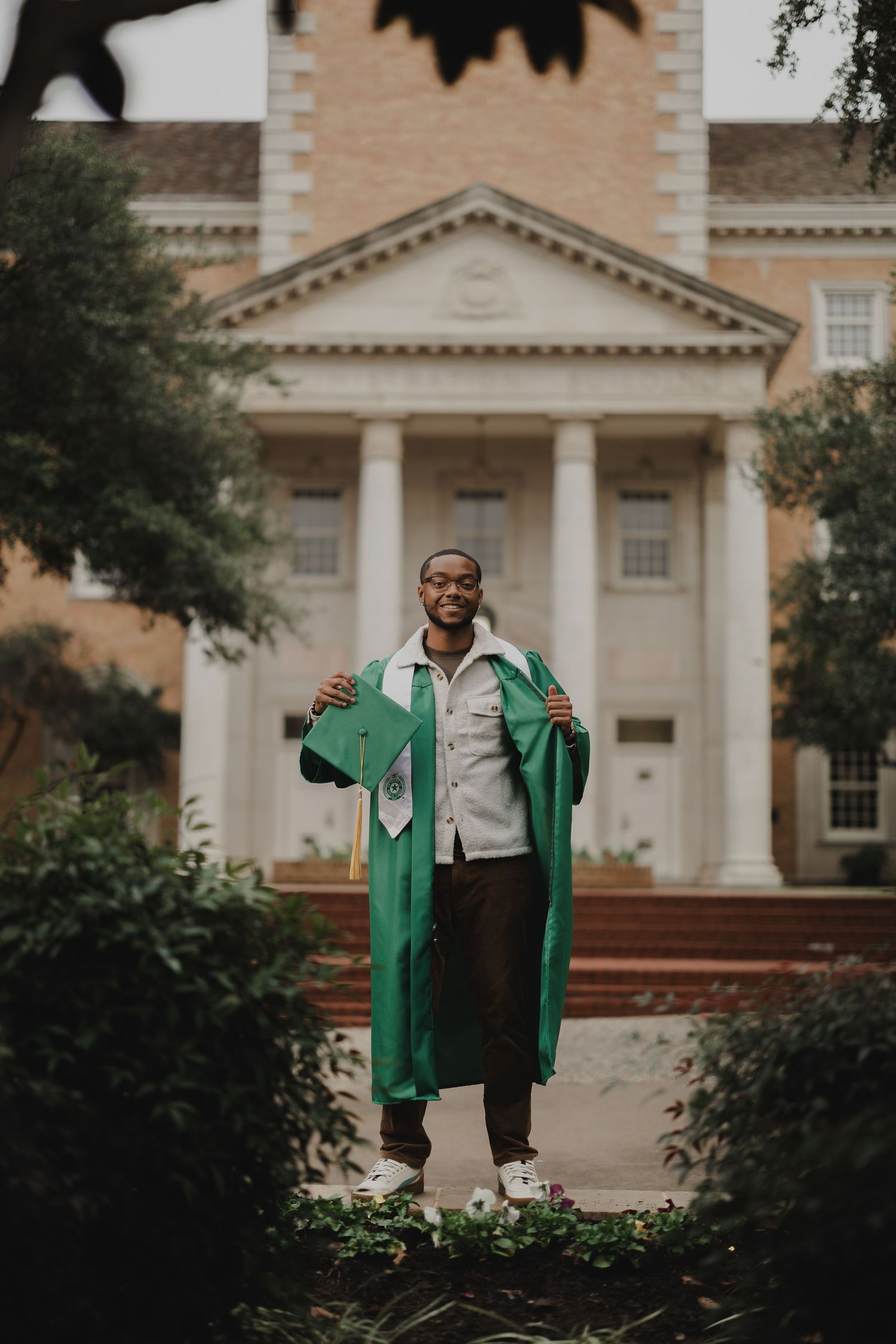 Graduate in green cap and gown standing in front of a stately colonial building with white columns.