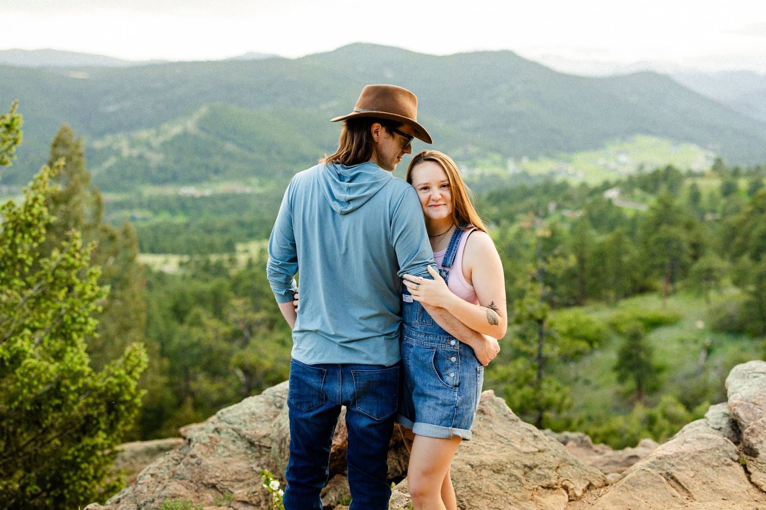 Couple standing on rocky overlook with scenic mountain valley and green forest views in background.