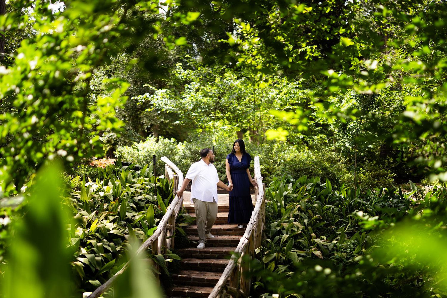 Couple holding hands walking up wooden stairs surrounded by lush green tropical foliage in a garden setting.